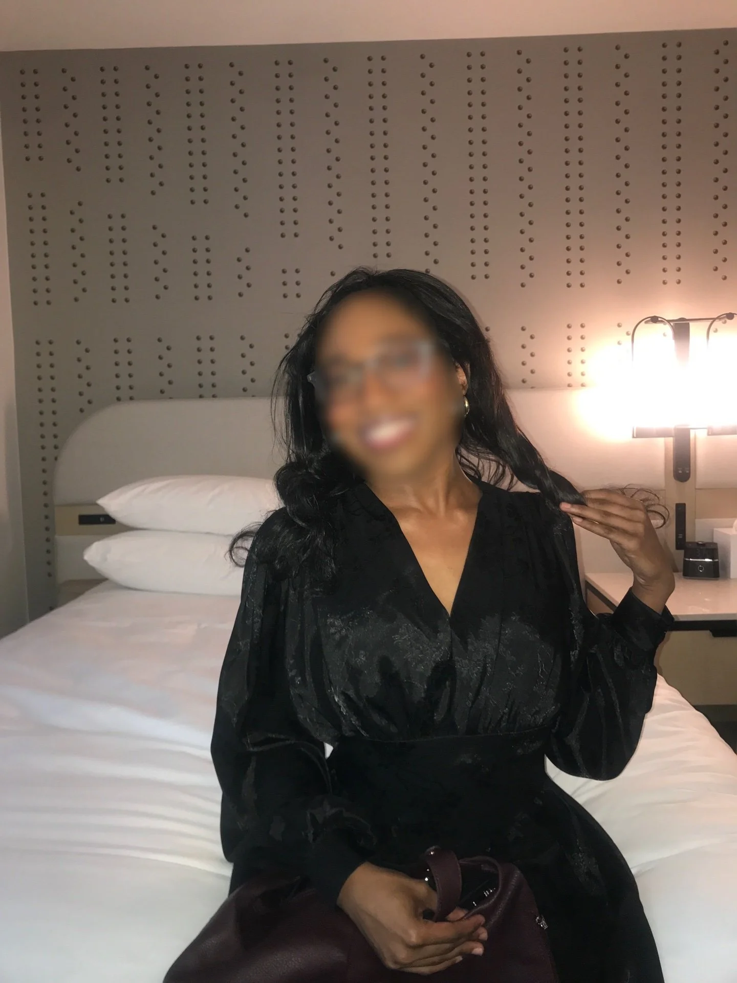 A woman with black hair and glasses sitting on a bed in a hotel room, smiling and looking at the camera. She is wearing a black dress and holding a maroon handbag.