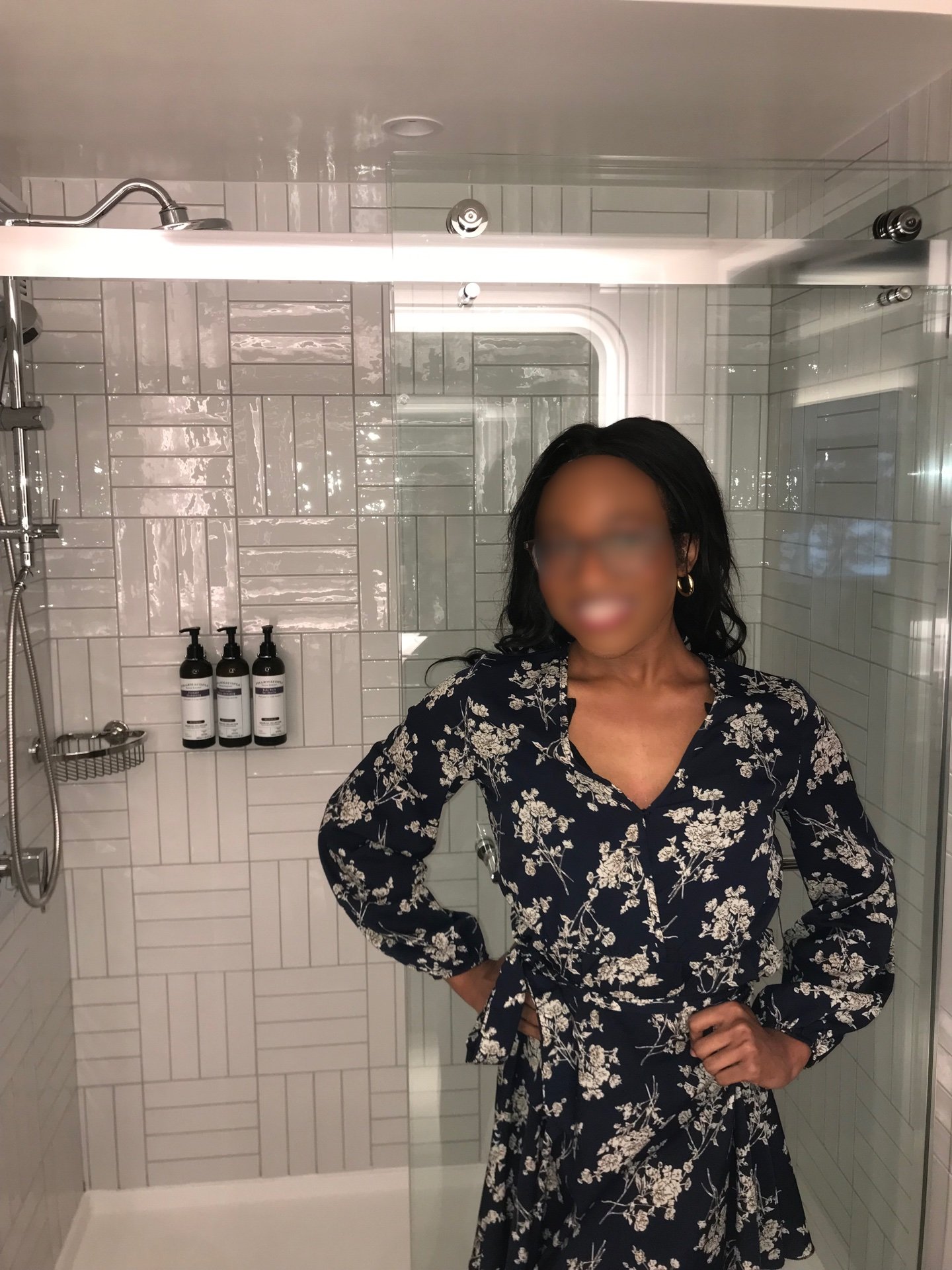 A woman standing in a glass-enclosed shower area, wearing a black dress with white floral patterns, posing with hands on hips.
