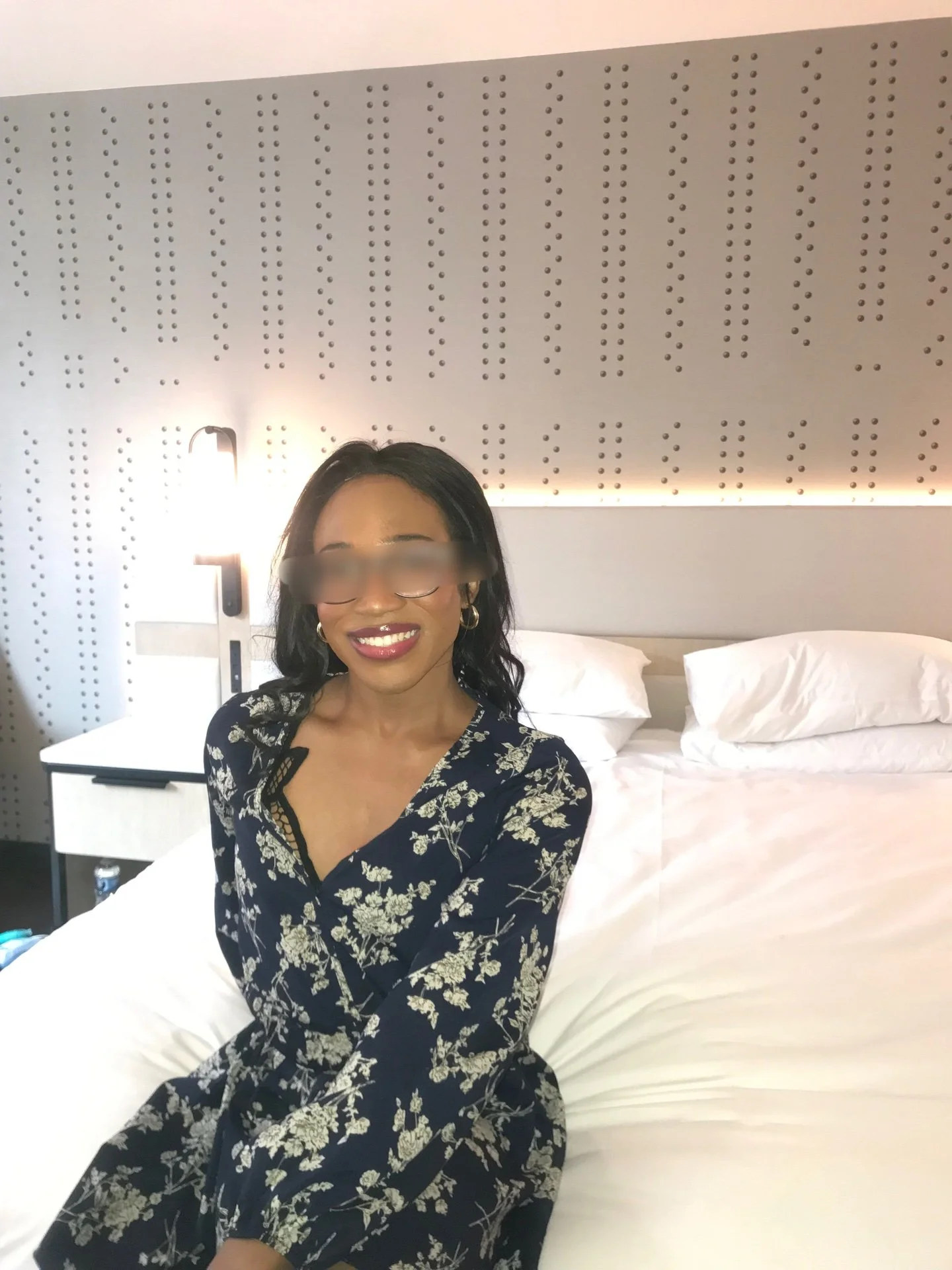A woman with dark, wavy hair and glasses, wearing a navy blue floral dress, is sitting on a bed in a modern hotel room with a textured gray headboard and white bedding, smiling at the camera.