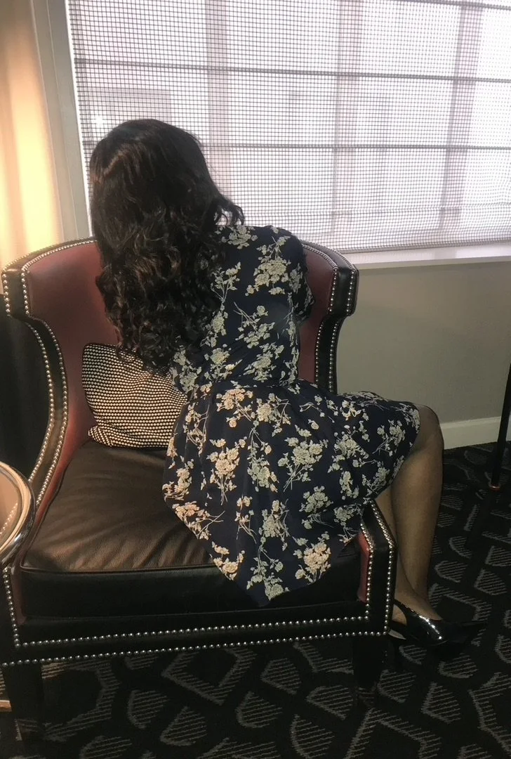 A woman with long, dark, curly hair sitting on a dark leather armchair, facing away from the camera, wearing a black dress with white floral print, black tights, and black shoes, in front of a window with a semi-transparent blind.