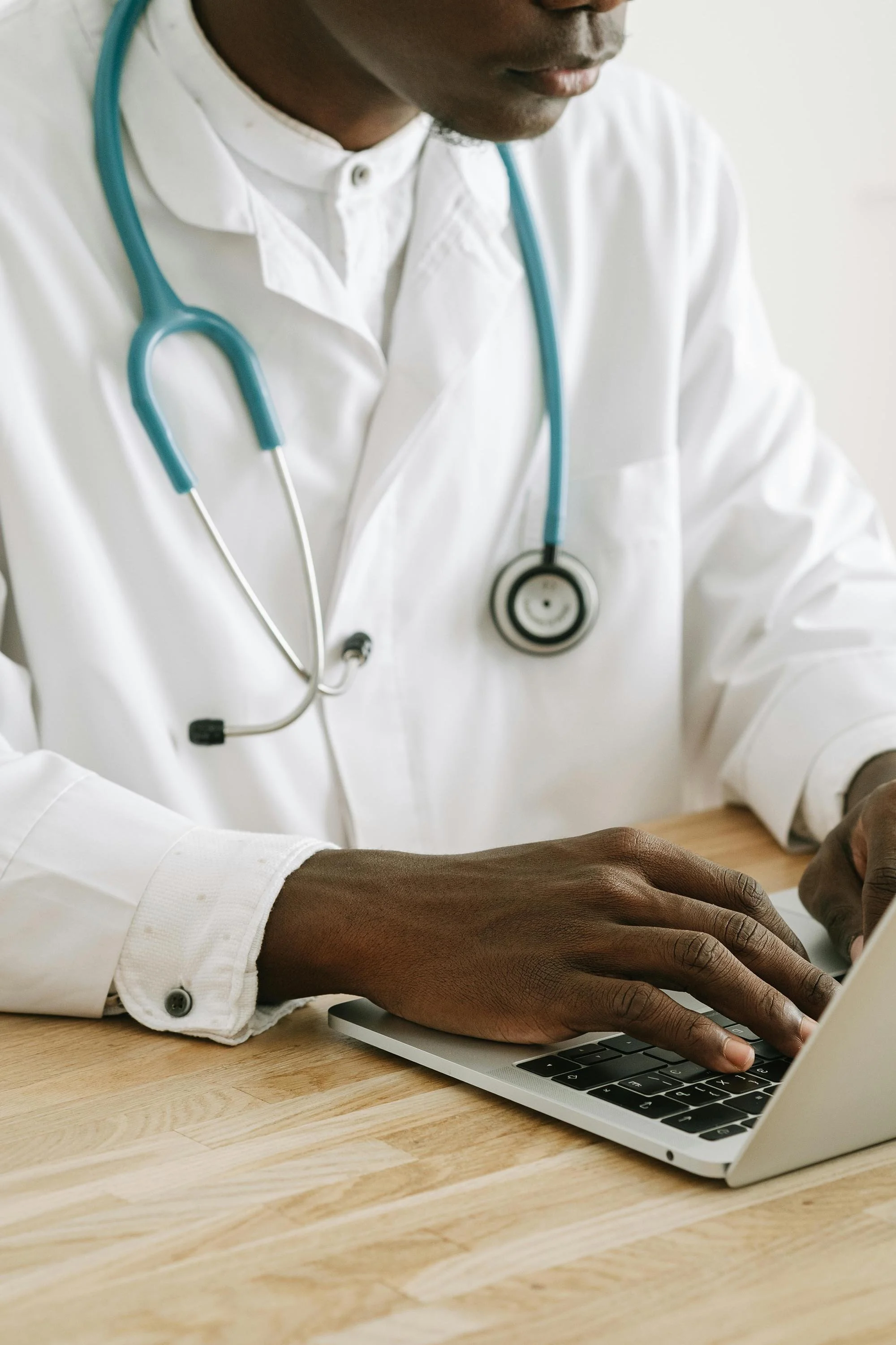 Close-up of a healthcare professional wearing a white coat and a stethoscope around their neck, typing on a laptop.