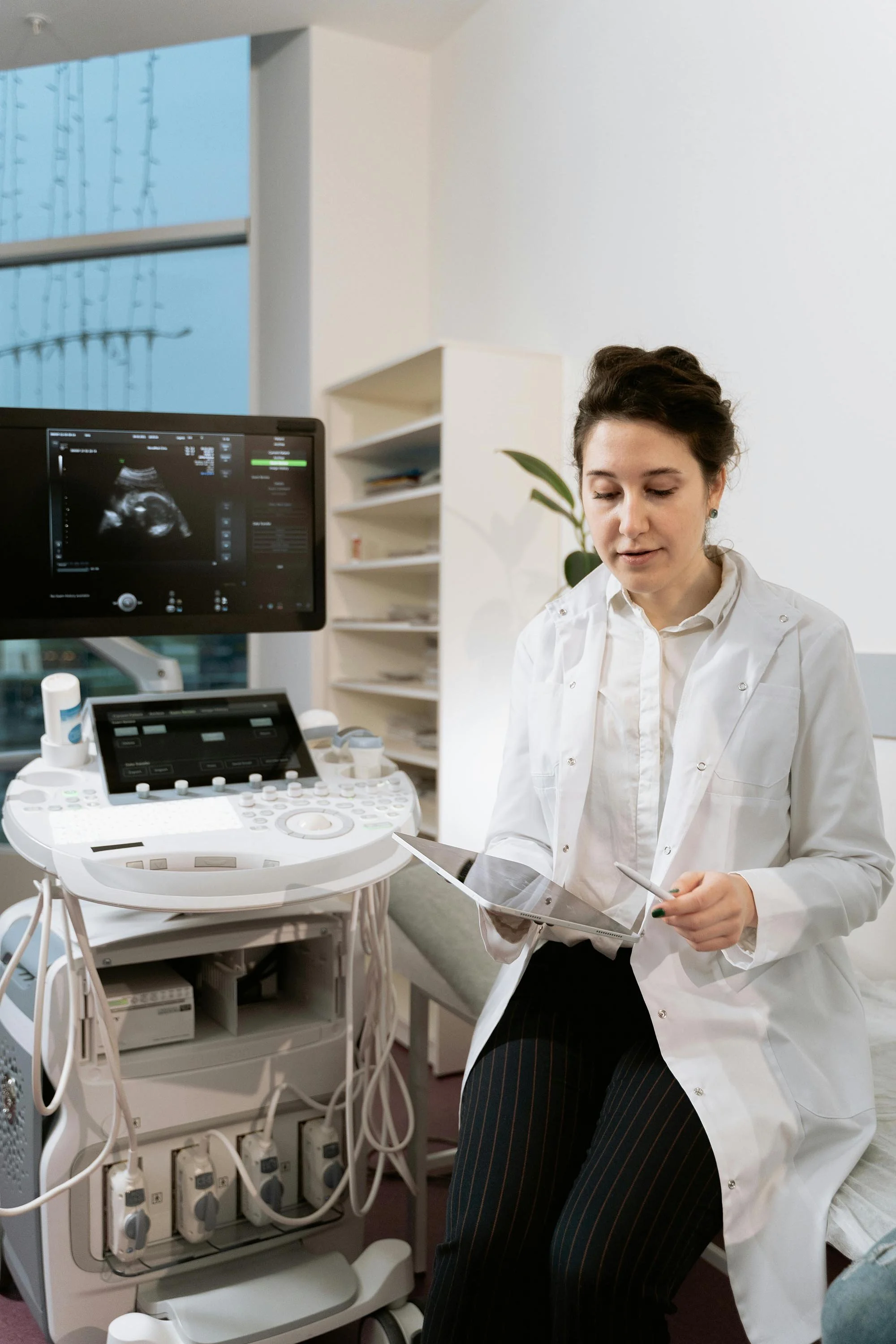 A female medical professional in a white lab coat sitting on an examination table, holding a tablet and examining ultrasound images. An ultrasound machine with a monitor showing an ultrasound scan is next to her in a clinical setting with white walls and shelves.