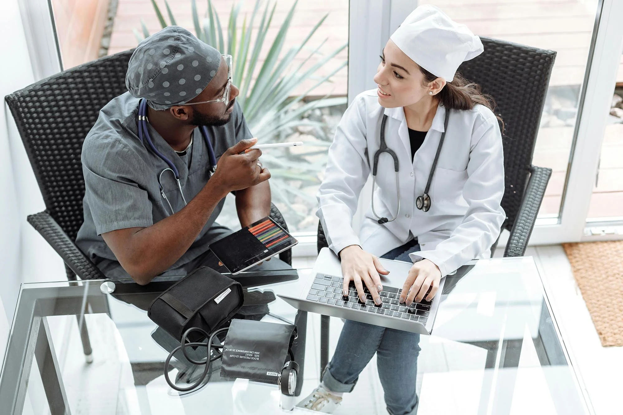 A healthcare professional, a woman in a white coat with a stethoscope, sitting at a glass table showing a laptop and medical supplies. She is talking with a male healthcare worker who is wearing scrubs, a surgical cap, and glasses, holding a color test strip chart and a pen. They are in a bright room with large windows.