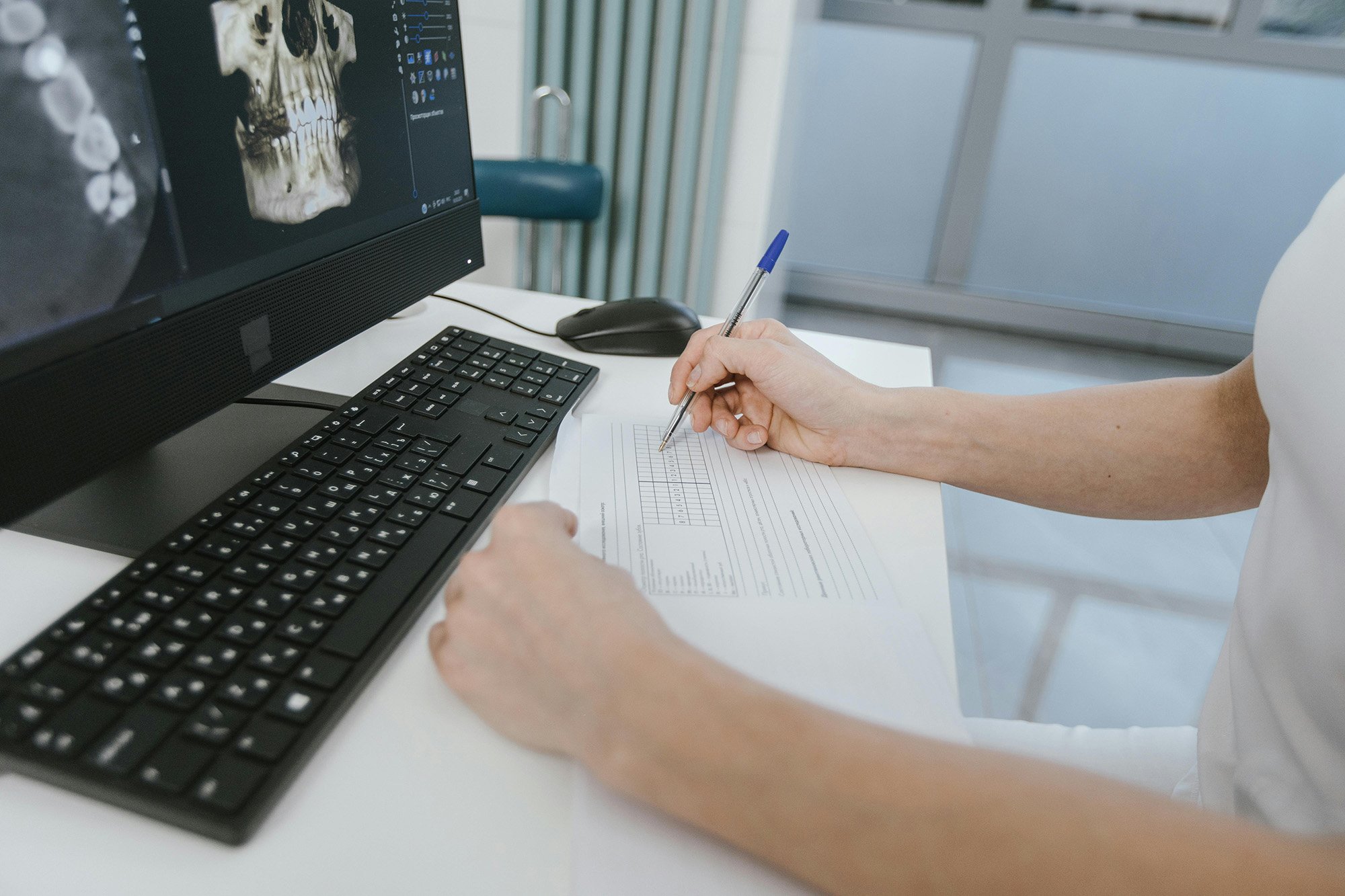 A person working at a desk with a computer monitor displaying a skull X-ray, writing on a document with a pen.