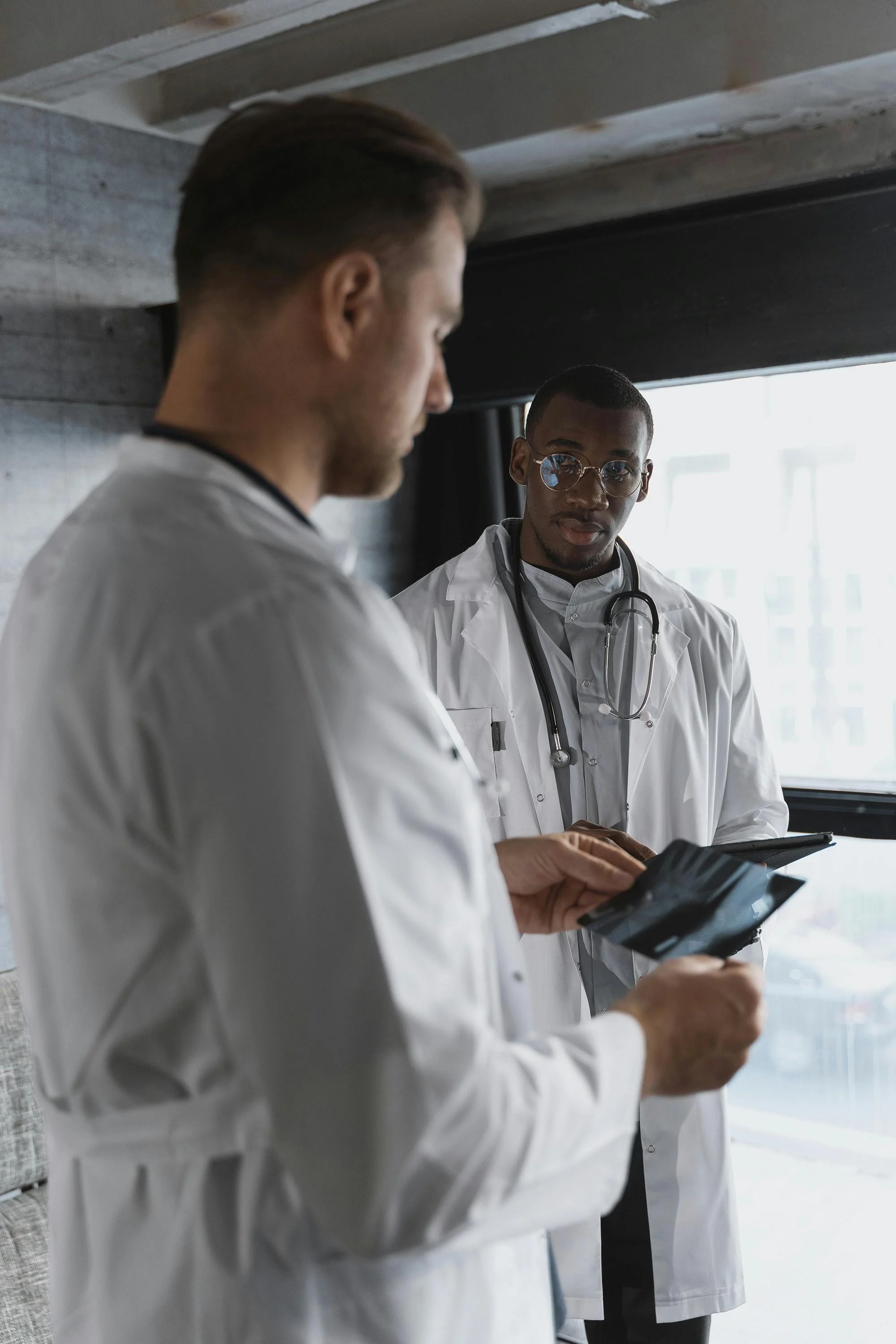 Two male doctors wearing white lab coats, one with a stethoscope around his neck, looking at medical images or tests inside a bright room near a window.