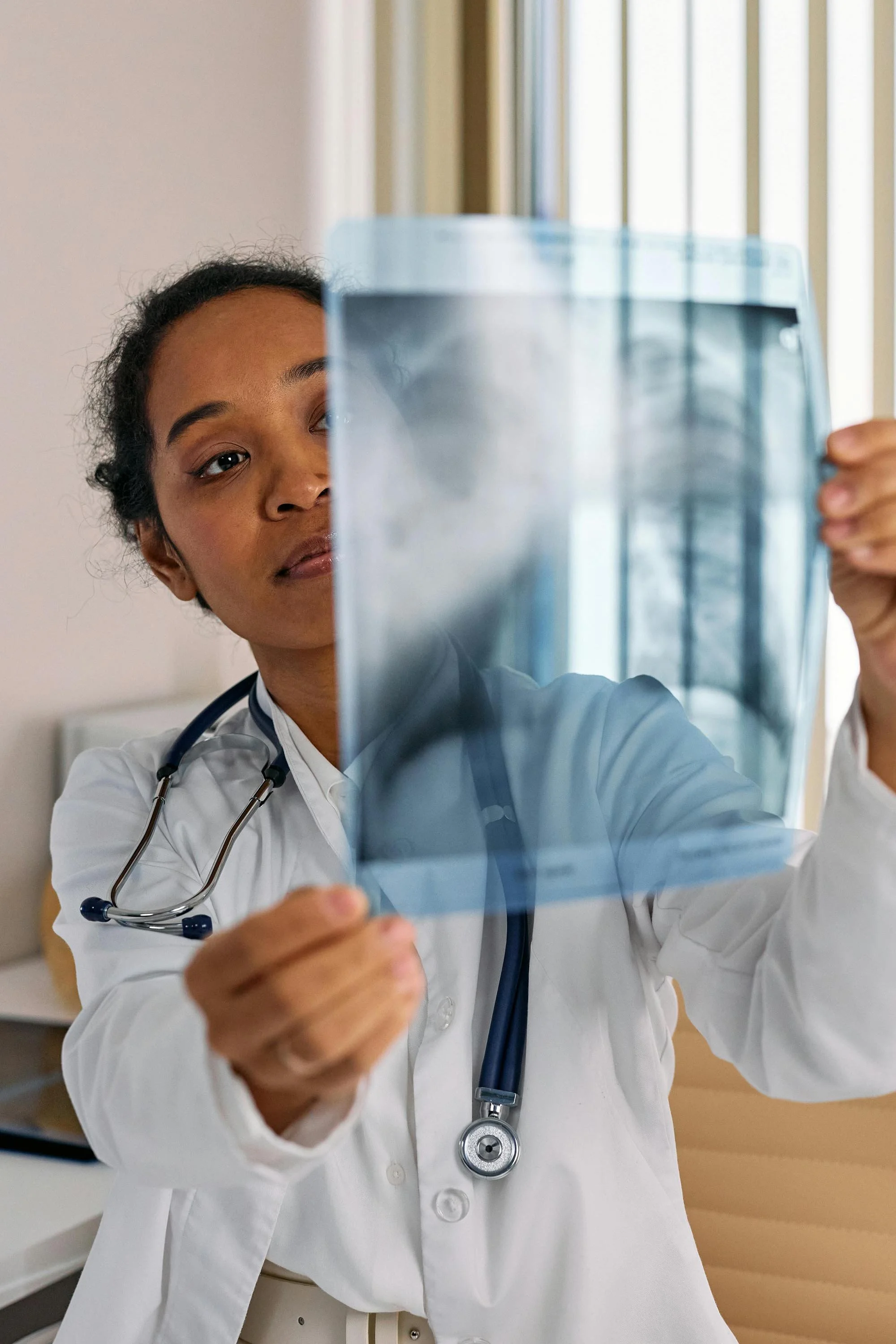 A female doctor wearing a white coat and stethoscope examining a chest X-ray film.