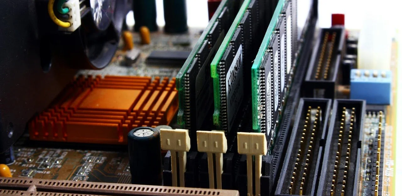 Close-up of a computer motherboard with multiple RAM sticks installed, featuring orange and green slots, and various electronic components.