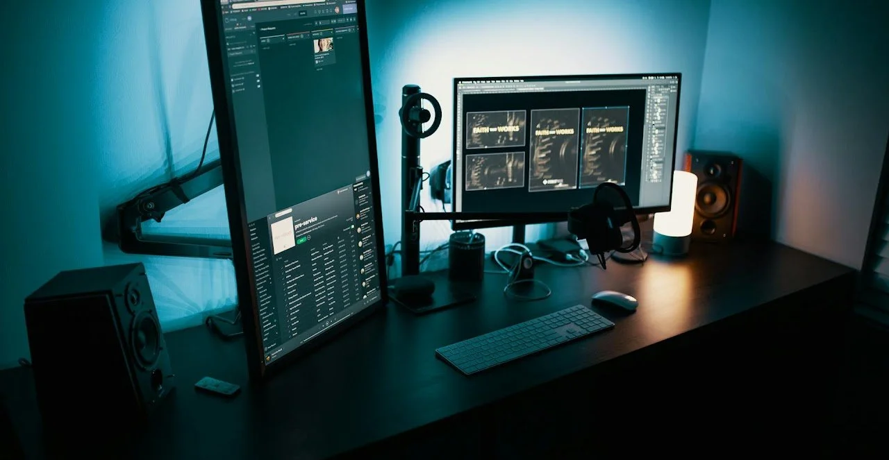 A dual-monitor computer setup on a dark desk with speakers, a keyboard, and a mouse, illuminated by a white lamp and a blue ambient light.