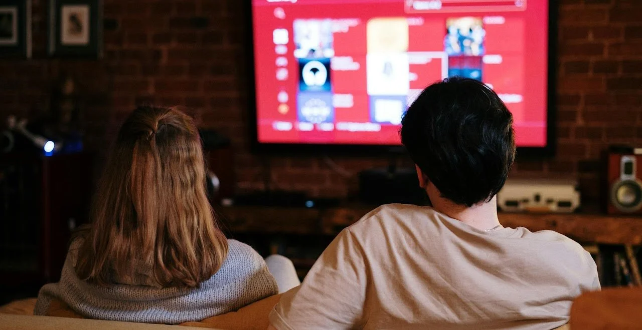 Two people sitting on a couch watching a television in a cozy living room with a brick wall, with a woman with brown hair and a man with black hair, facing away from the camera.