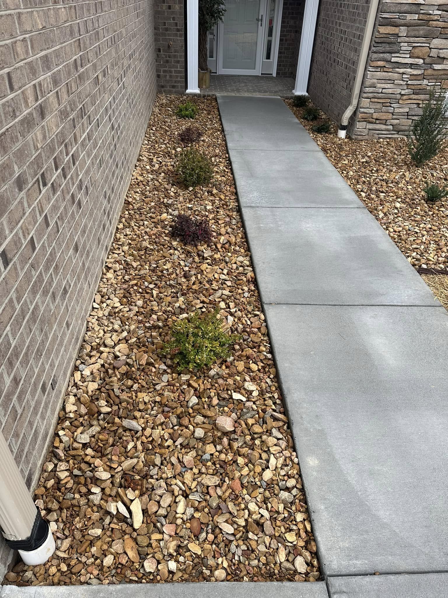 Sidewalk leading to house entrance with small plants and rocks along the side, brick and stone exterior walls, and a front door visible in the background.
