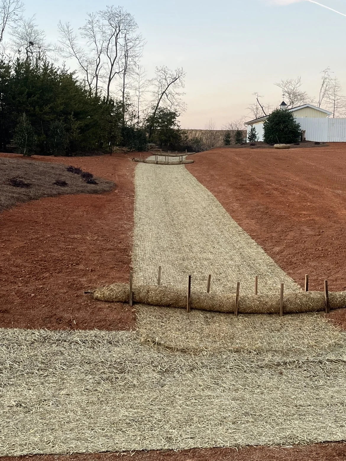 A pathway made of straw or similar material in a rural setting, bordered by stakes and wooden posts, leading towards a white building with a white fence and leafless trees in the background.