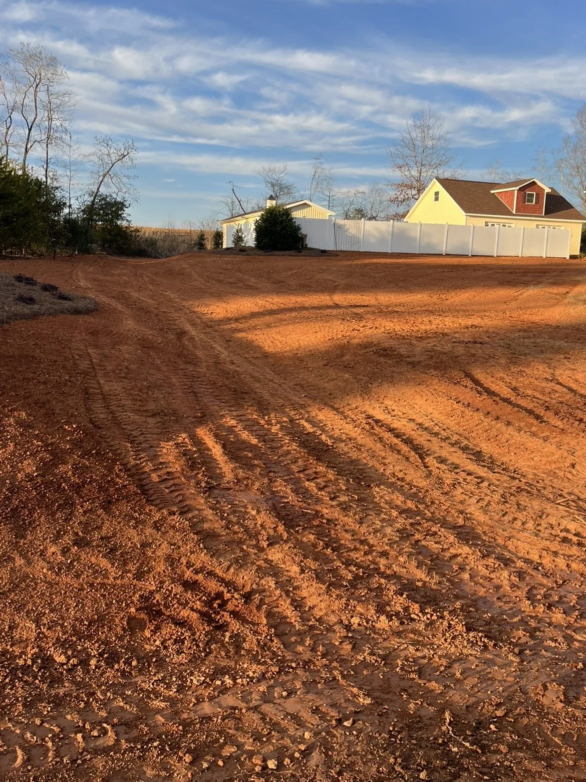 A dirt driveway with tire tracks, leading to a backyard with a yellow house, a white fence, and leafless trees under a blue sky with some clouds.
