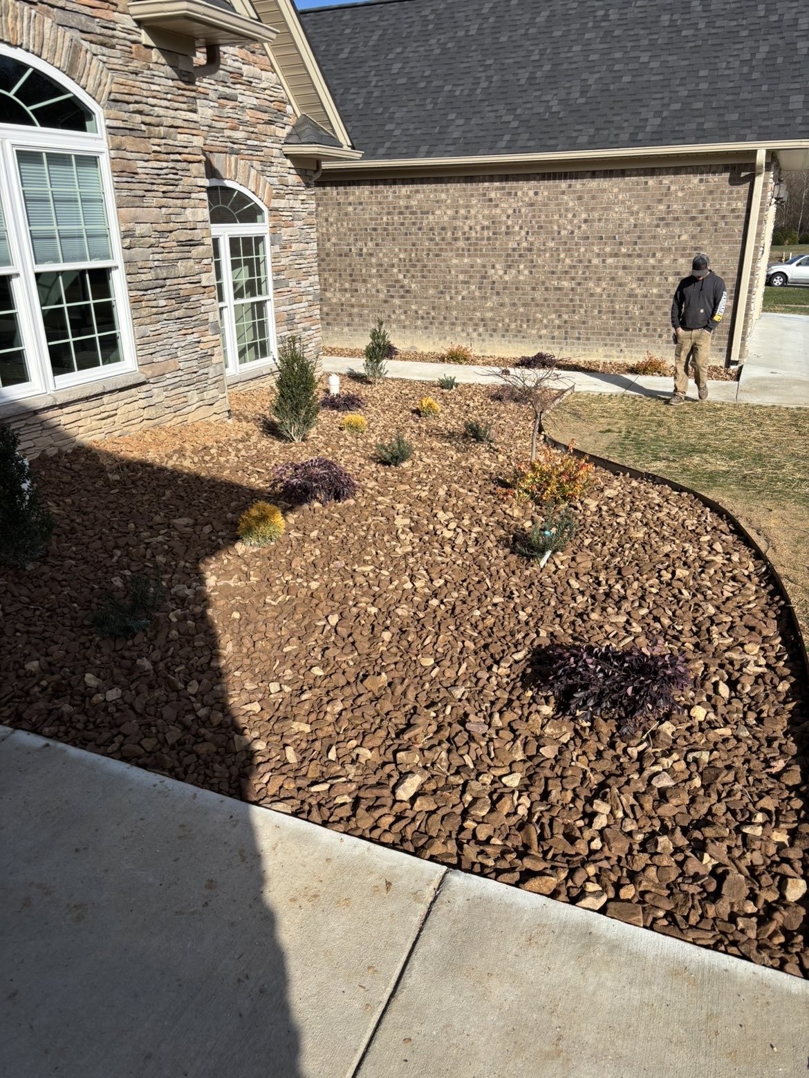 A house with stone and brick exterior walls, large windows, and a sloped roof. The front yard has a landscaped garden bed covered with reddish-brown rocks, containing small shrubs and plants. A person in dark clothing and a cap is walking on the sidewalk near the driveway.