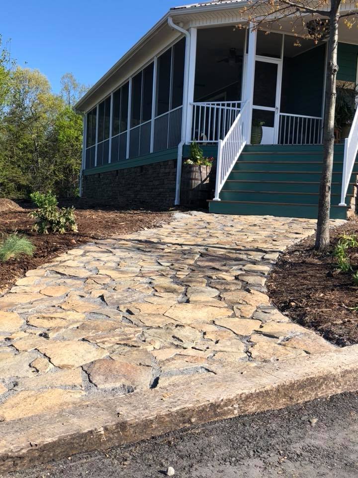Stone pathway leading to a screened porch of a house with a staircase, railing, and a tree nearby.