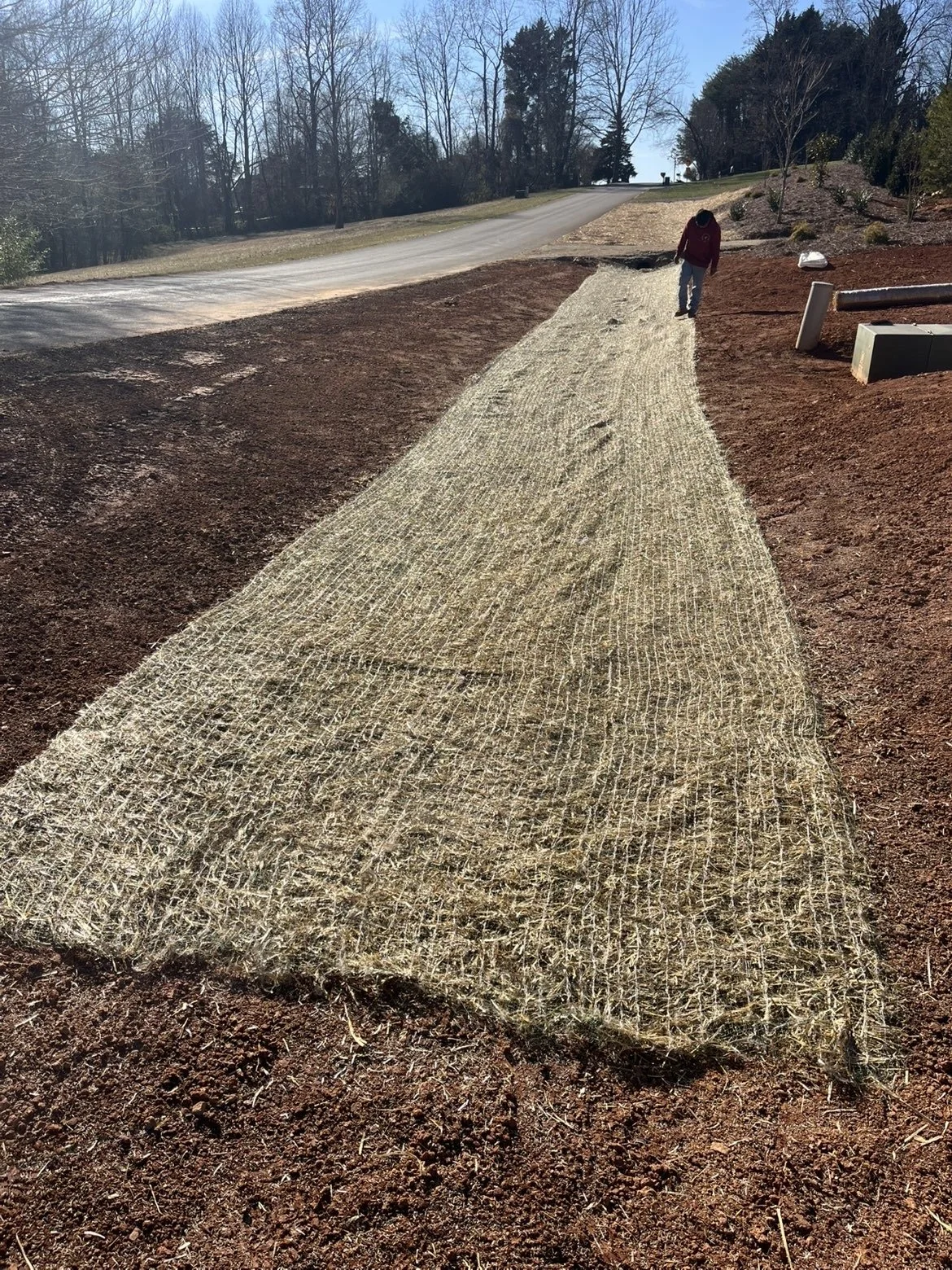 A person walking on a newly laid gravel pathway on a sloped terrain surrounded by bare trees and some equipment on the side.