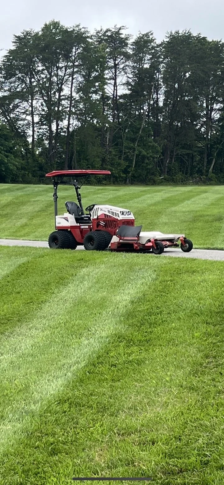 Image of a red and white riding lawn mower parked on a concrete path in a lush green park or golf course, with trees and grass in the background.