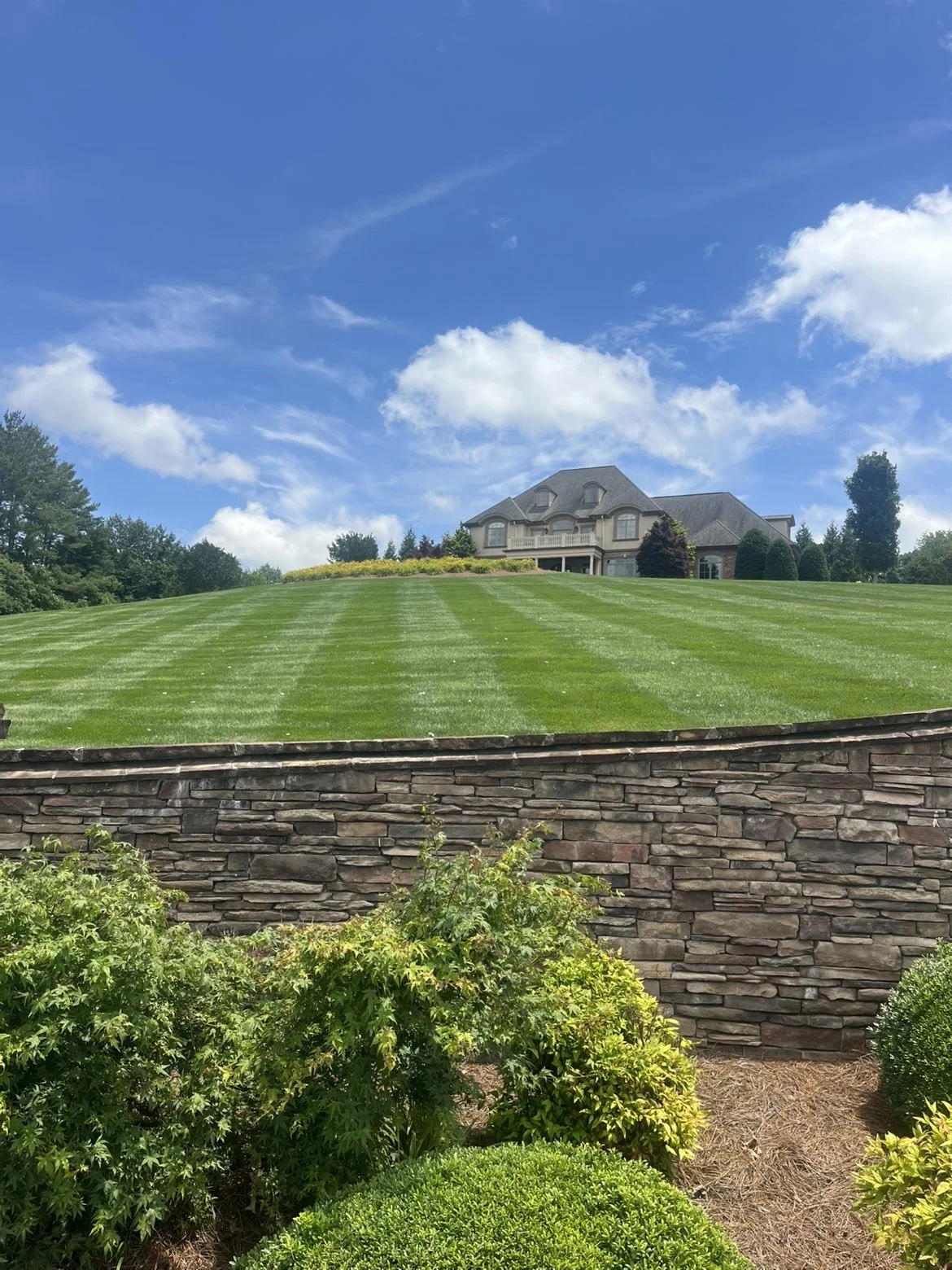 Large house on a hill with a well-maintained lawn, stone retaining wall, and landscaped bushes in the foreground under a blue sky with white clouds.