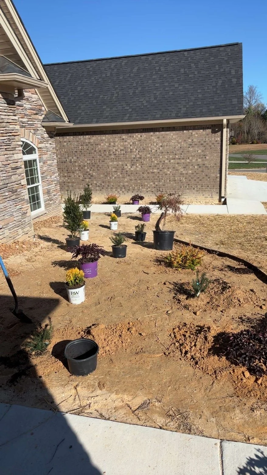 A backyard garden with potted plants and small shrubs, next to a brick house with stone accents. The soil is bare, and the garden is in early stages of planting.