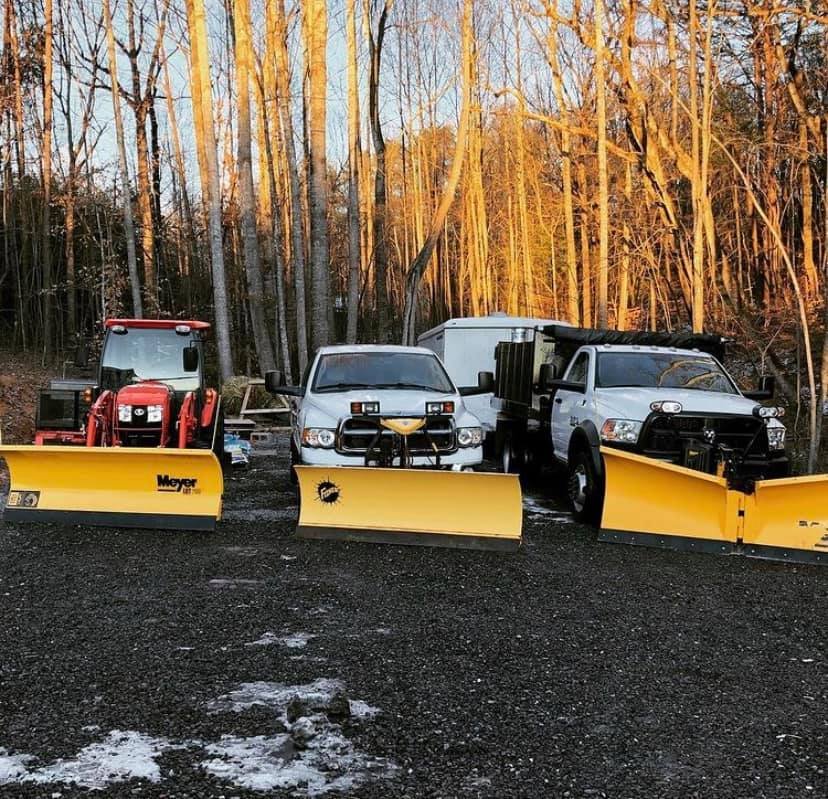 Three vehicles equipped for snow removal with yellow snowplow blades attached, parked in front of leafless trees during winter.