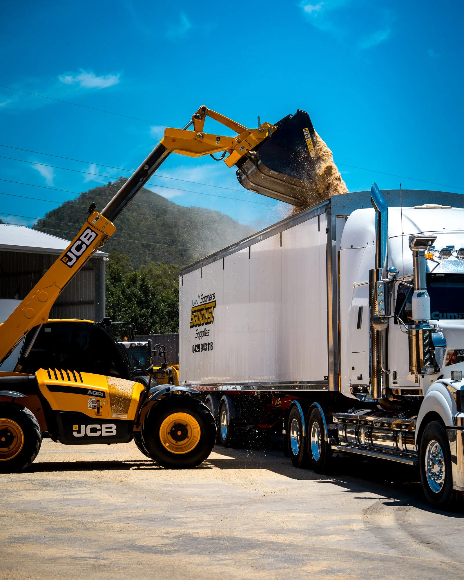 A JCB front loader with a yellow arm and black bucket loading sand or gravel into a large white truck with a silver cab on a construction or industrial site, blue sky in the background.
