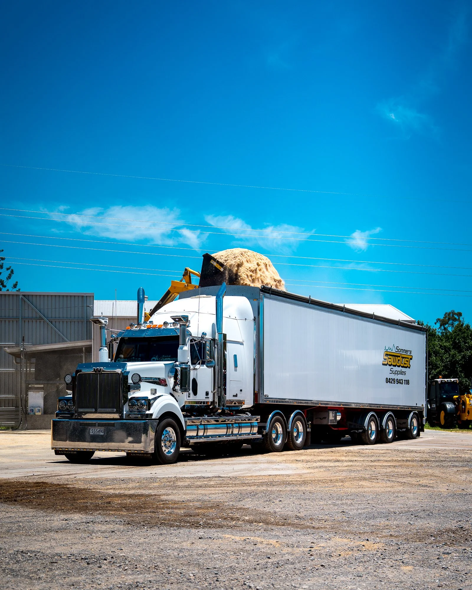 A large white semi-truck with a trailer, parked on a dirt lot, with a yellow excavator loader nearby. The truck has a black grill and chrome details, and the trailer has logo and contact information for a supplier.