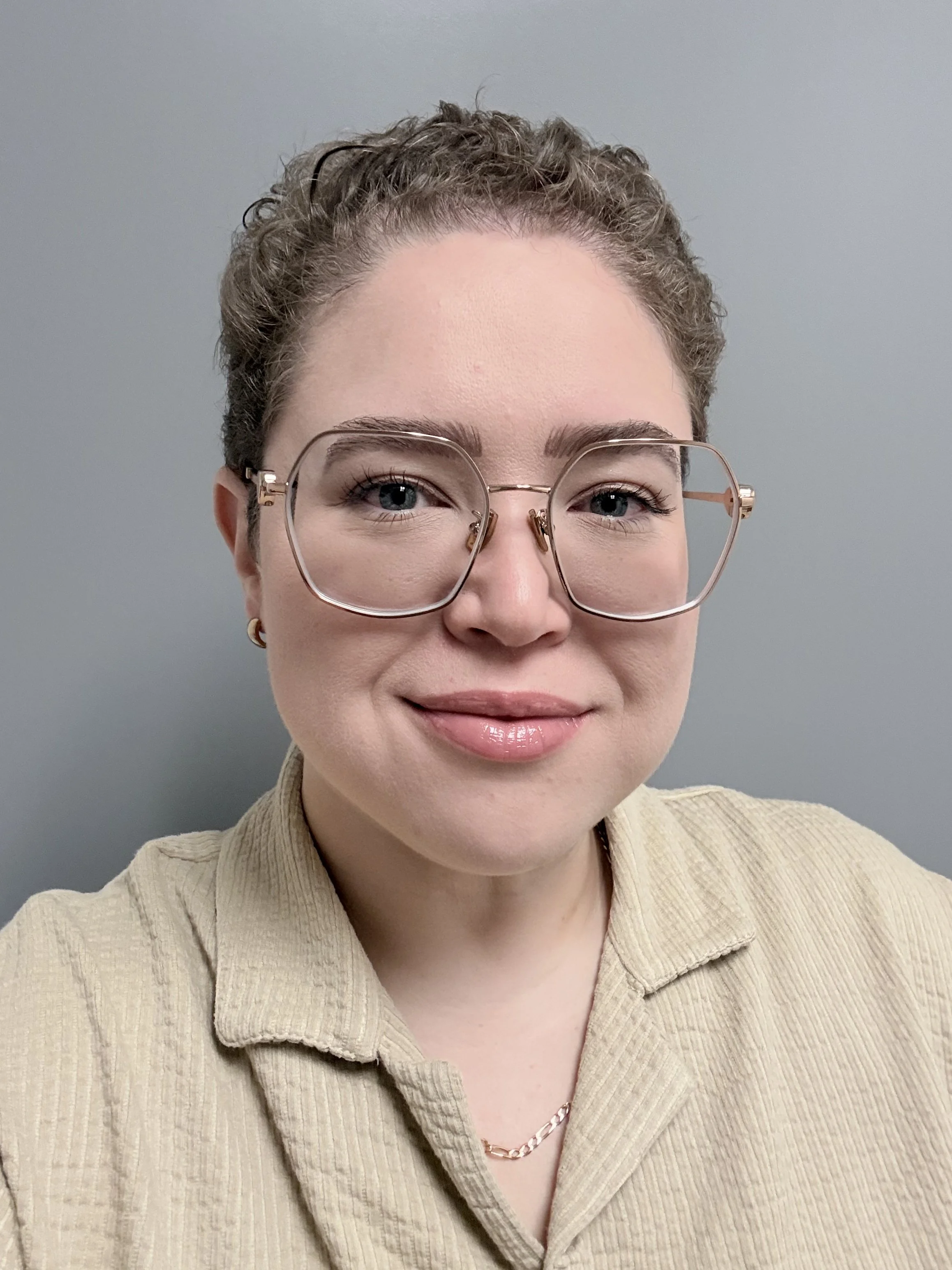 Close-up of a woman with short curly hair, wearing large clear glasses, a beige collared shirt, and a chain necklace, smiling against a plain gray background.