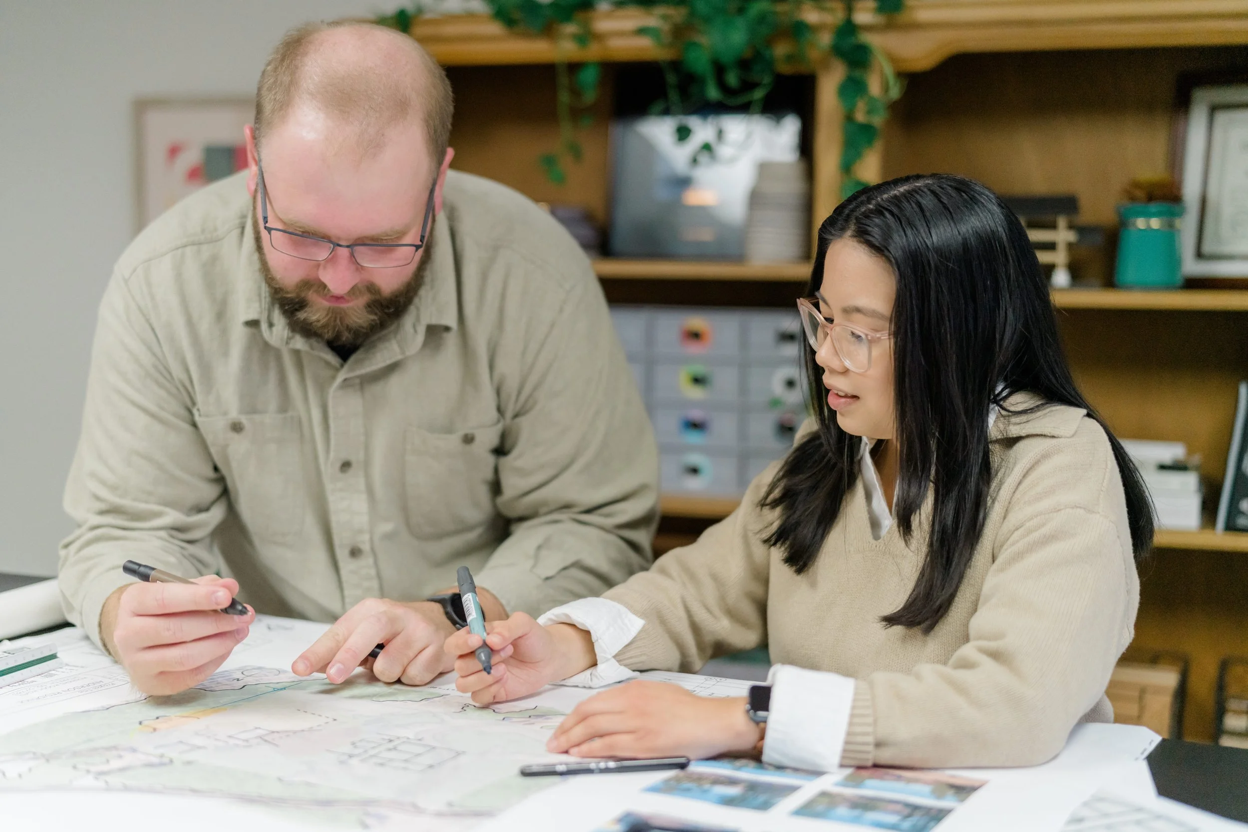 Two people, a man and a woman, are collaborating on a project at a table. The man is pointing at a map or blueprint, and the woman is holding a marker, with photos and documents spread out on the table.