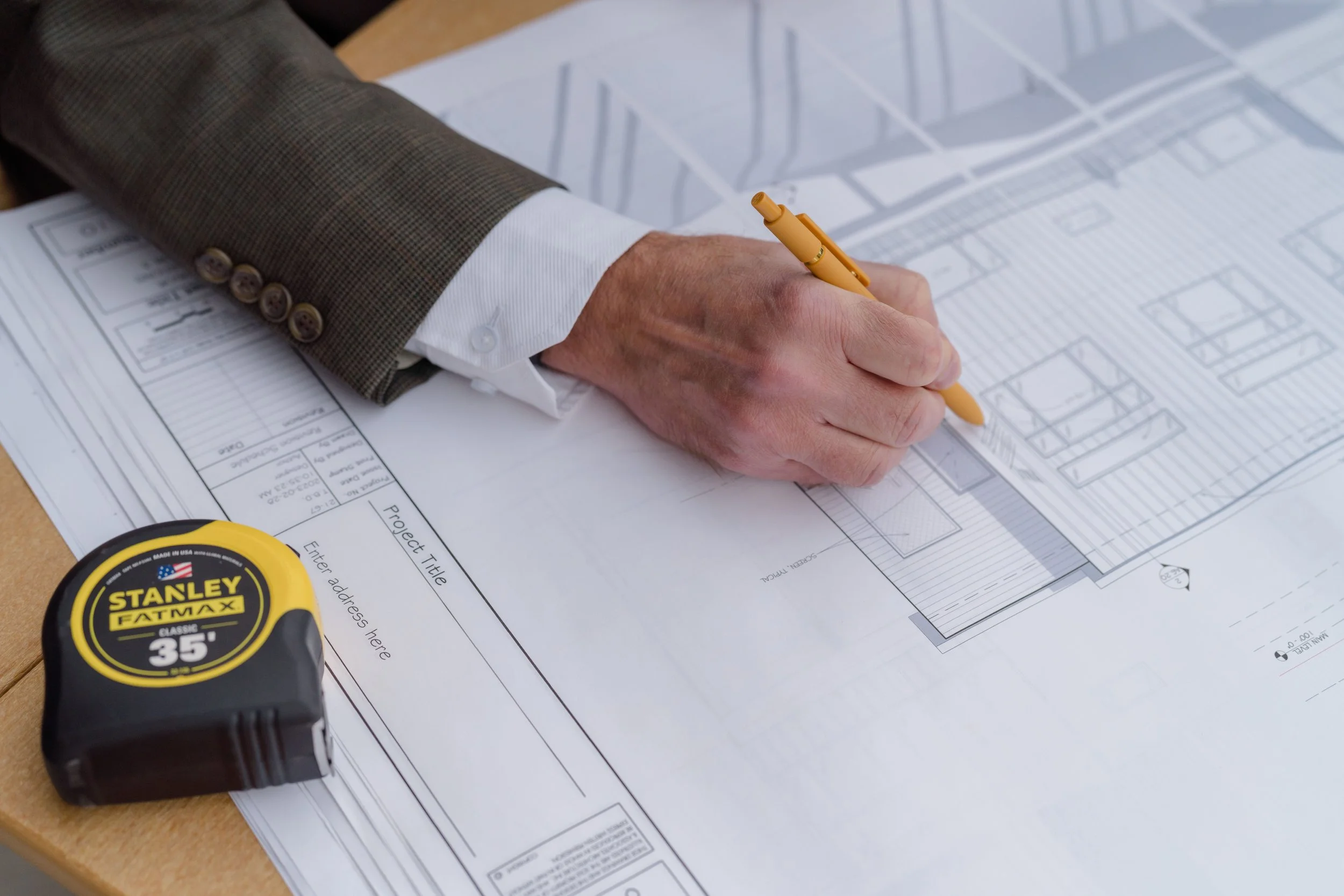A person in a suit is drawing or reviewing architectural blueprints on a table, with a yellow Stanley FATMAX tape measure nearby.