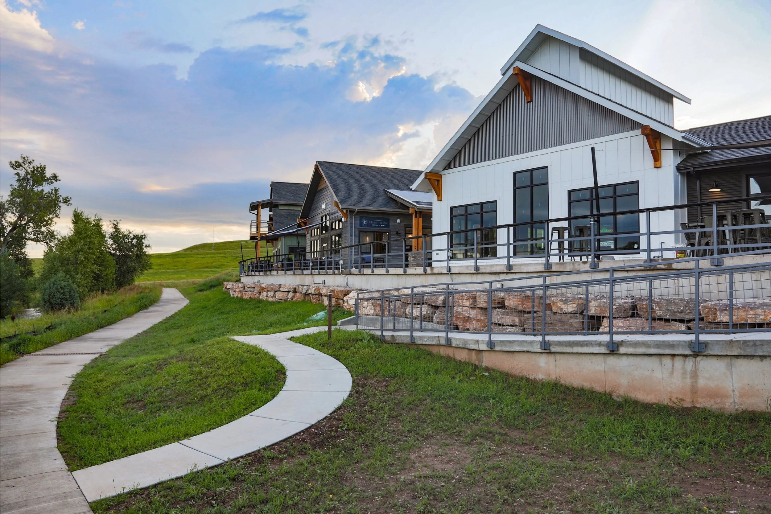A modern building with large windows and a balcony, situated on a grassy hillside, with a winding concrete pathway leading up to it. The sky is partly cloudy and the area has trees and green landscape.