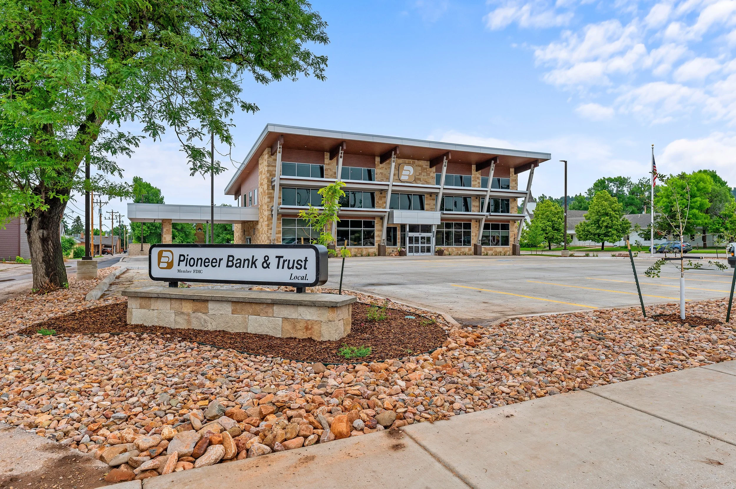 Modern commercial building with a sign for Pioneer Bank & Trust in a landscaped area with rocks and trees, featuring a mostly empty parking lot under a blue sky with some clouds.
