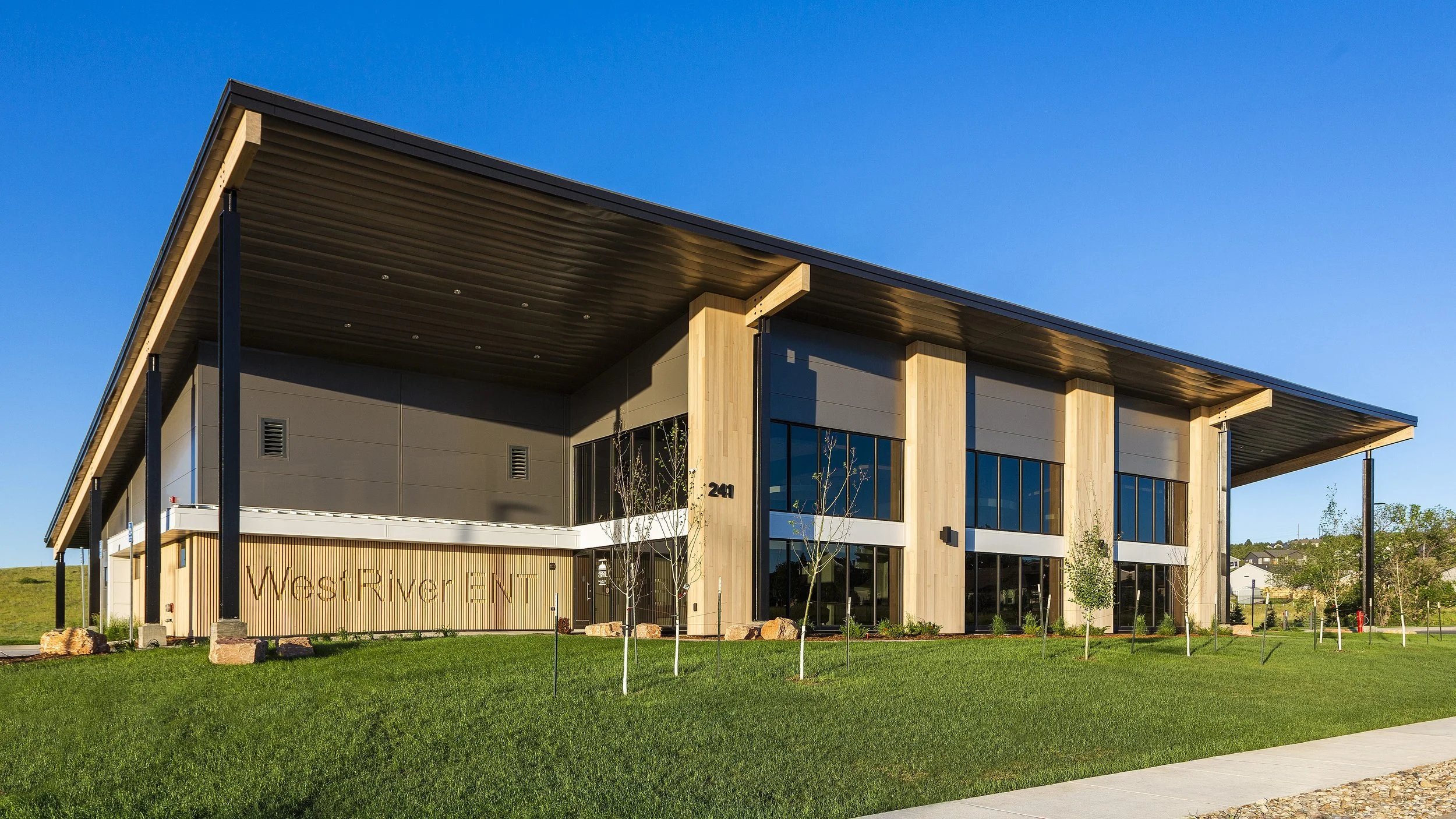 Modern two-story building with large glass windows, wooden accents, a sloped roof, and a landscaped lawn in front, under a clear blue sky.