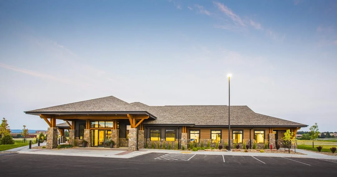 A modern, single-story building with stone and wood exterior, large windows, and a parking lot in front.
