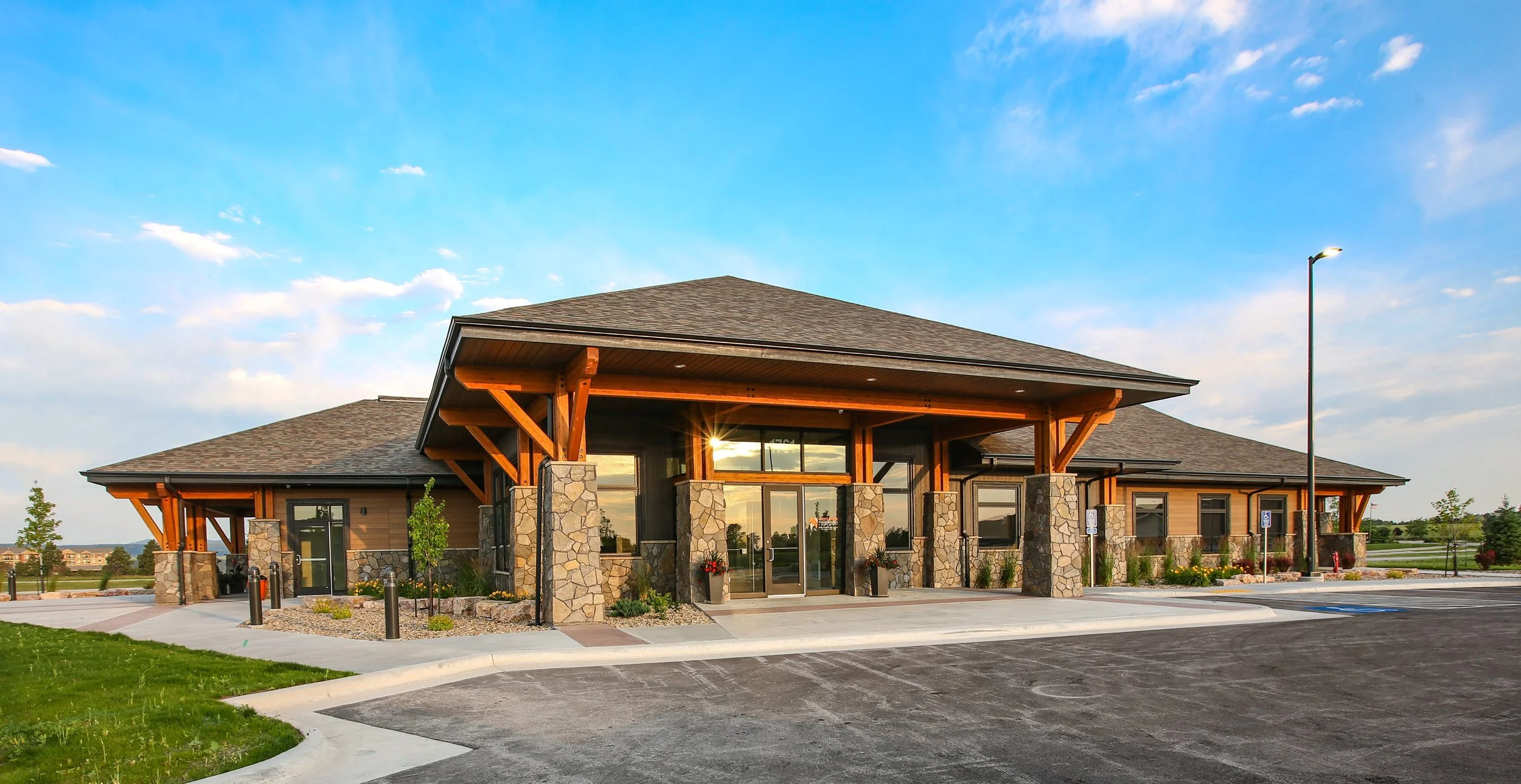 Modern building with stone and wood exterior, large glass doors, and a pitched roof, surrounded by a parking lot and landscaped greenery under a blue sky with clouds.