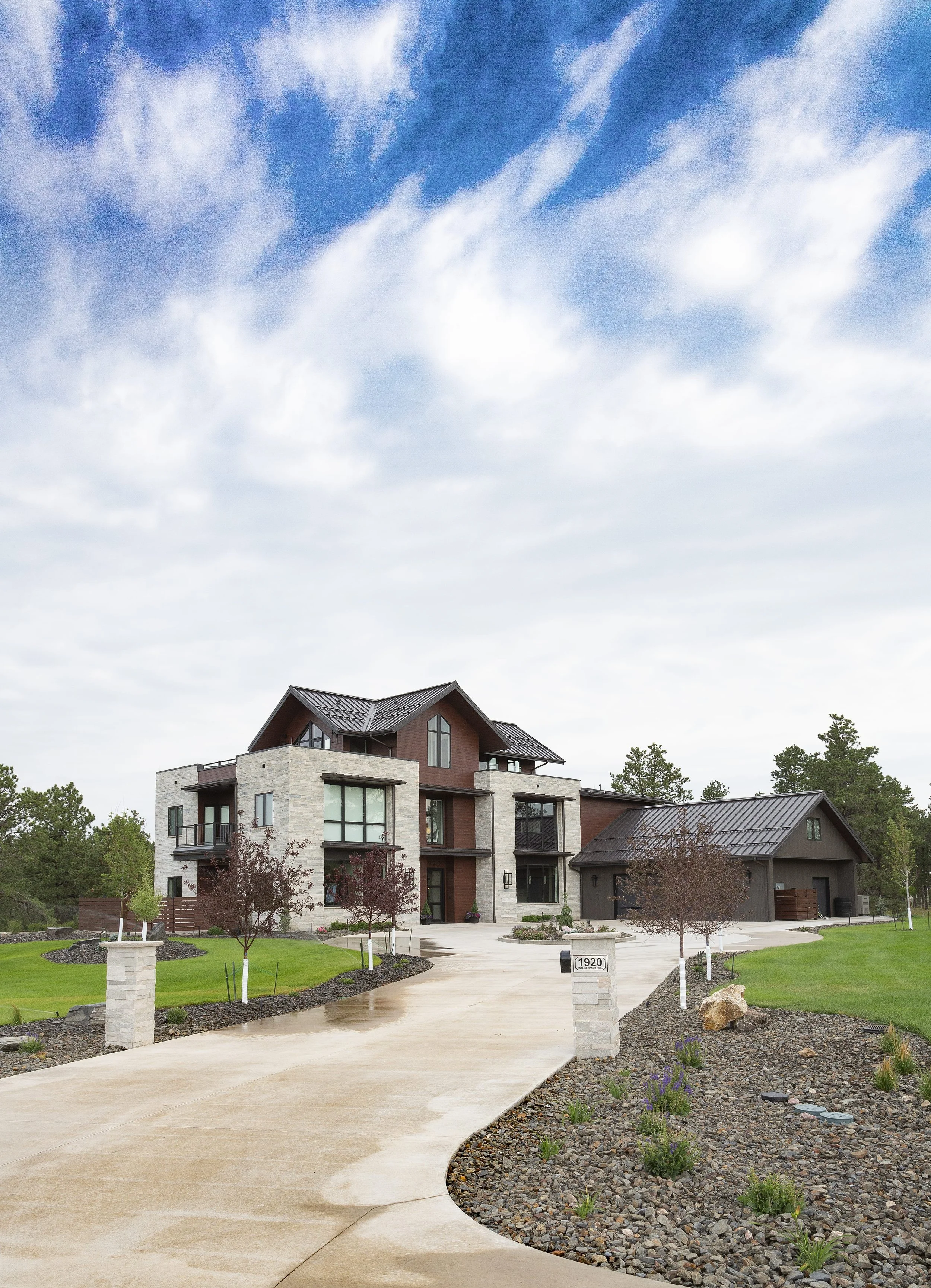 Modern house with stone and wood exterior, surrounded by landscaped yard and driveway, under a cloudy sky.