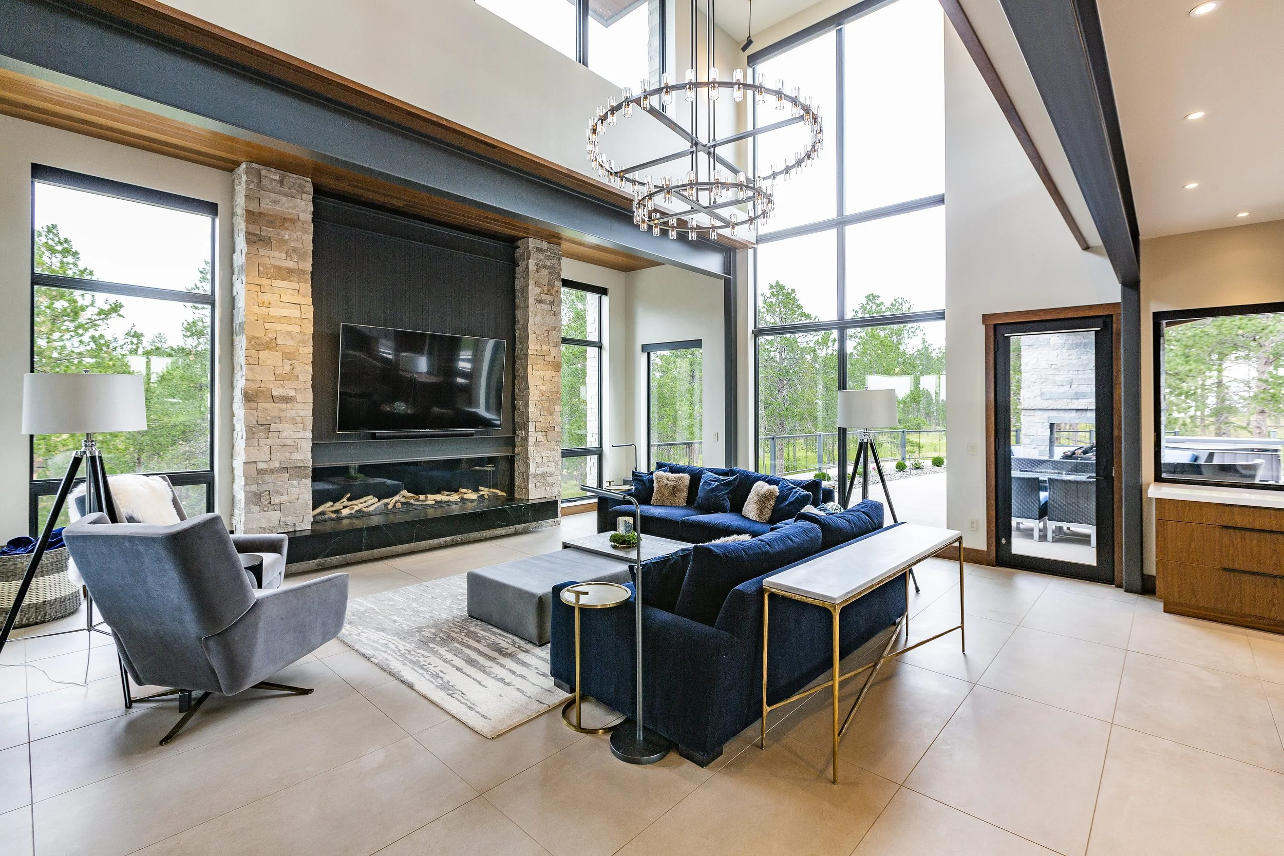 Modern living room with large windows, a stone fireplace with a mounted TV, a blue sectional sofa, a gray armchair, and a glass chandelier.