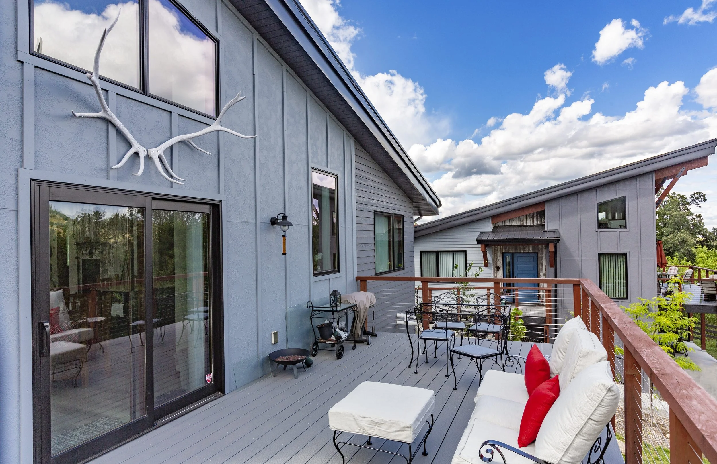 Outdoor balcony with white and red cushions on a white sofa, a small round ottoman, metal chairs around a table, a grill, and a railing with a view of neighboring houses and trees, under a partly cloudy sky.