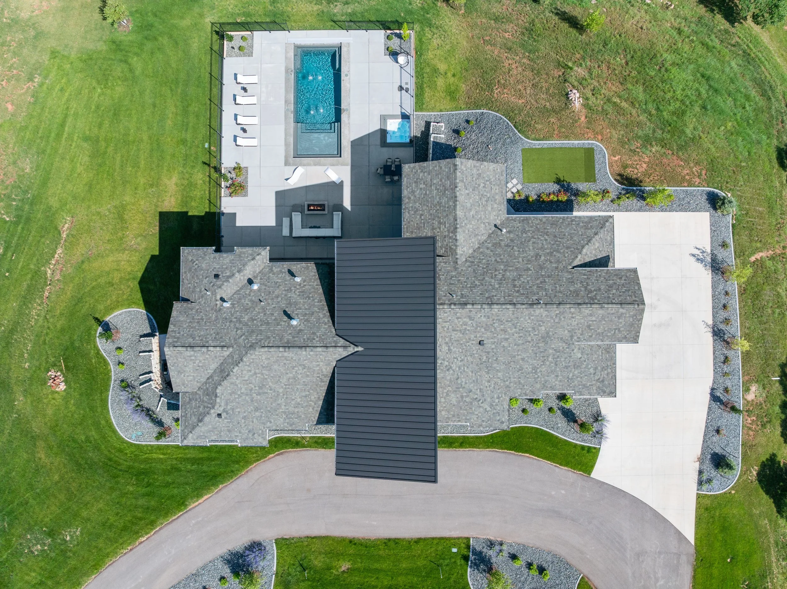 Aerial view of a house with a gray shingled roof, a large concrete driveway, a backyard pool area with lounge chairs, a fire pit, and landscaping, surrounded by green lawns and trees.