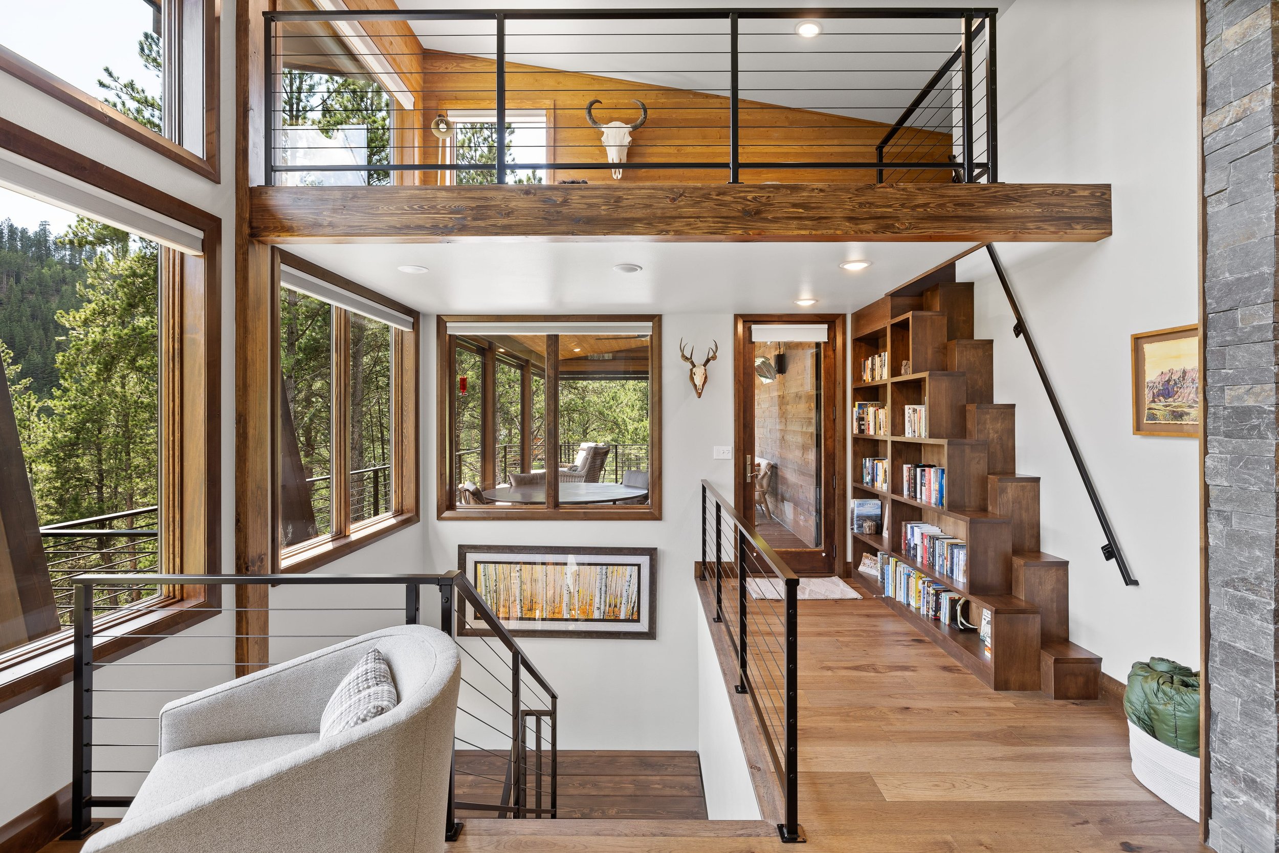 Interior view of a modern house with large windows, wooden floors, a staircase with a bookshelf, a balcony with a deer skull mounted on the wall, and trees visible outside.