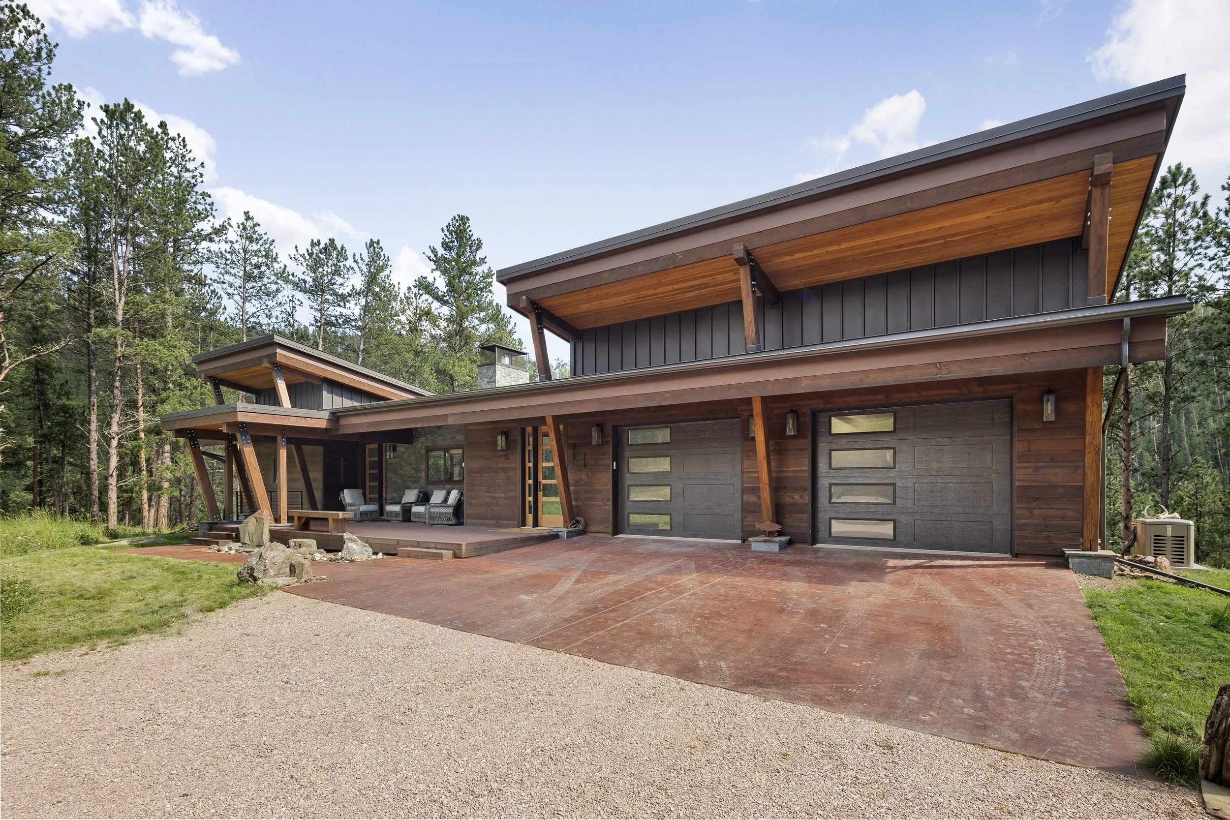 Modern house surrounded by trees, featuring a large driveway, wooden exterior, and outdoor seating area.