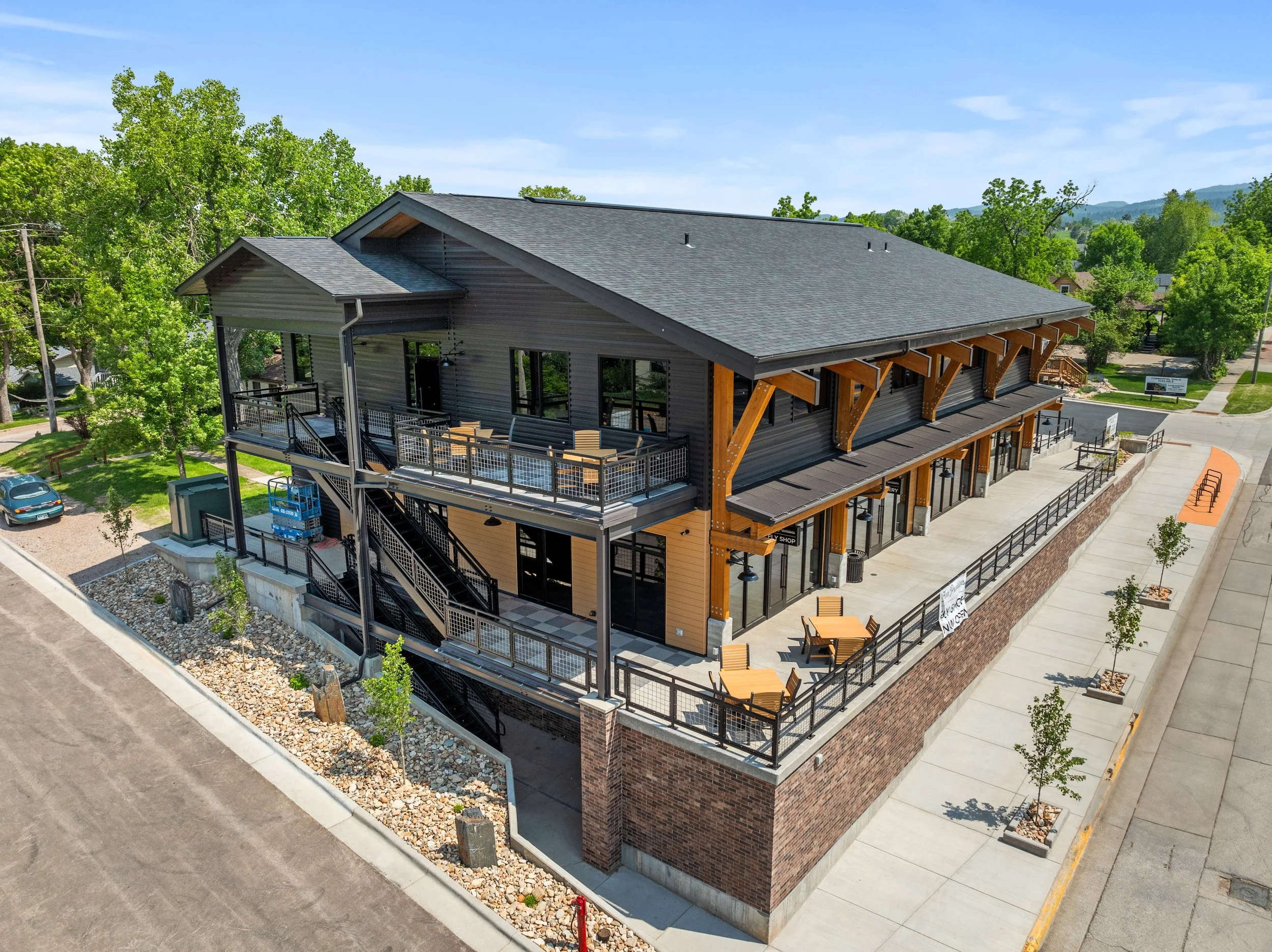 A modern two-story commercial building with outdoor seating on a terrace and sidewalk, surrounded by trees, with a parking lot and street in the background.