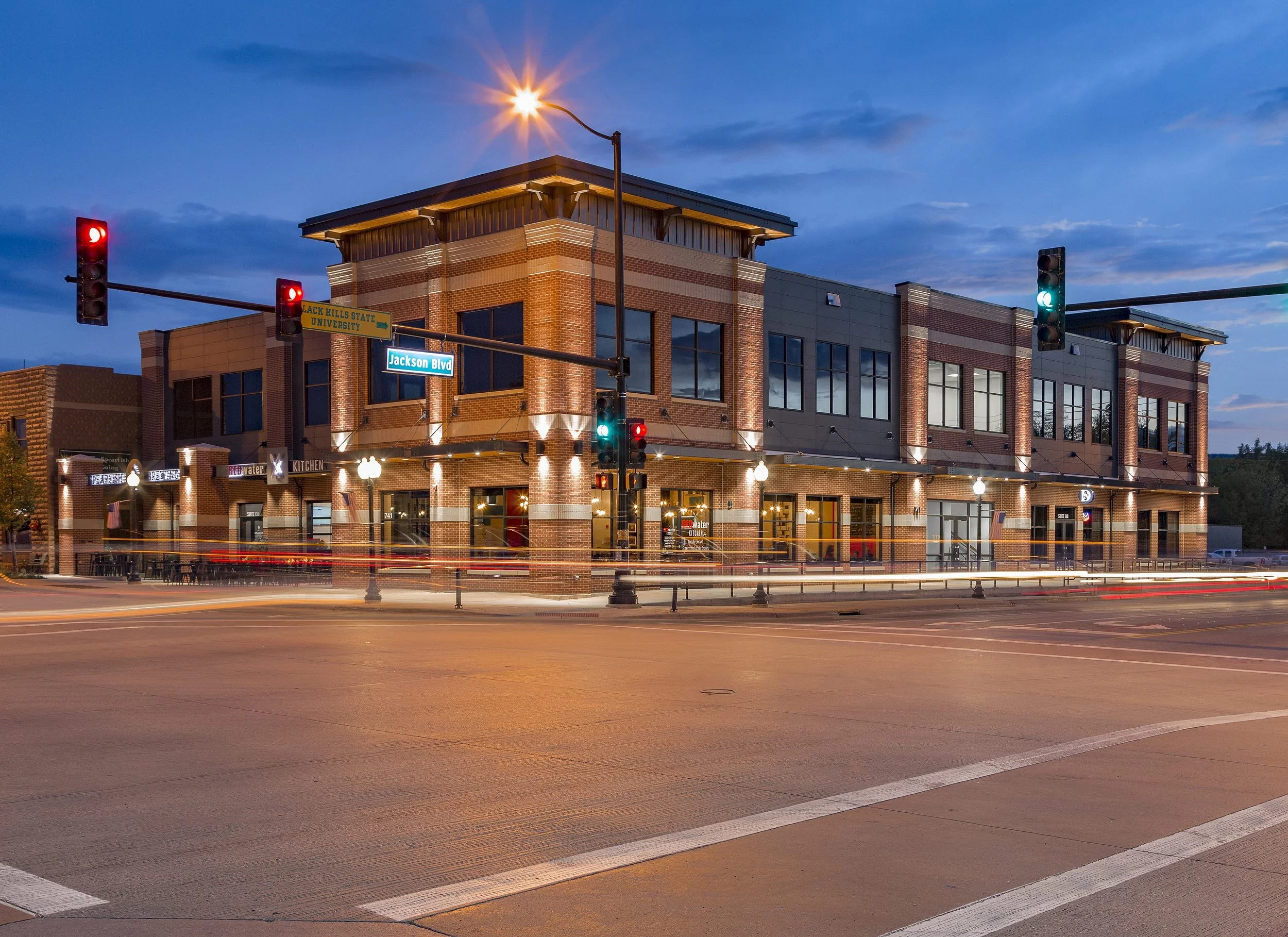 A two-story commercial building at the corner of a street with traffic lights and street signs, illuminated at dusk.