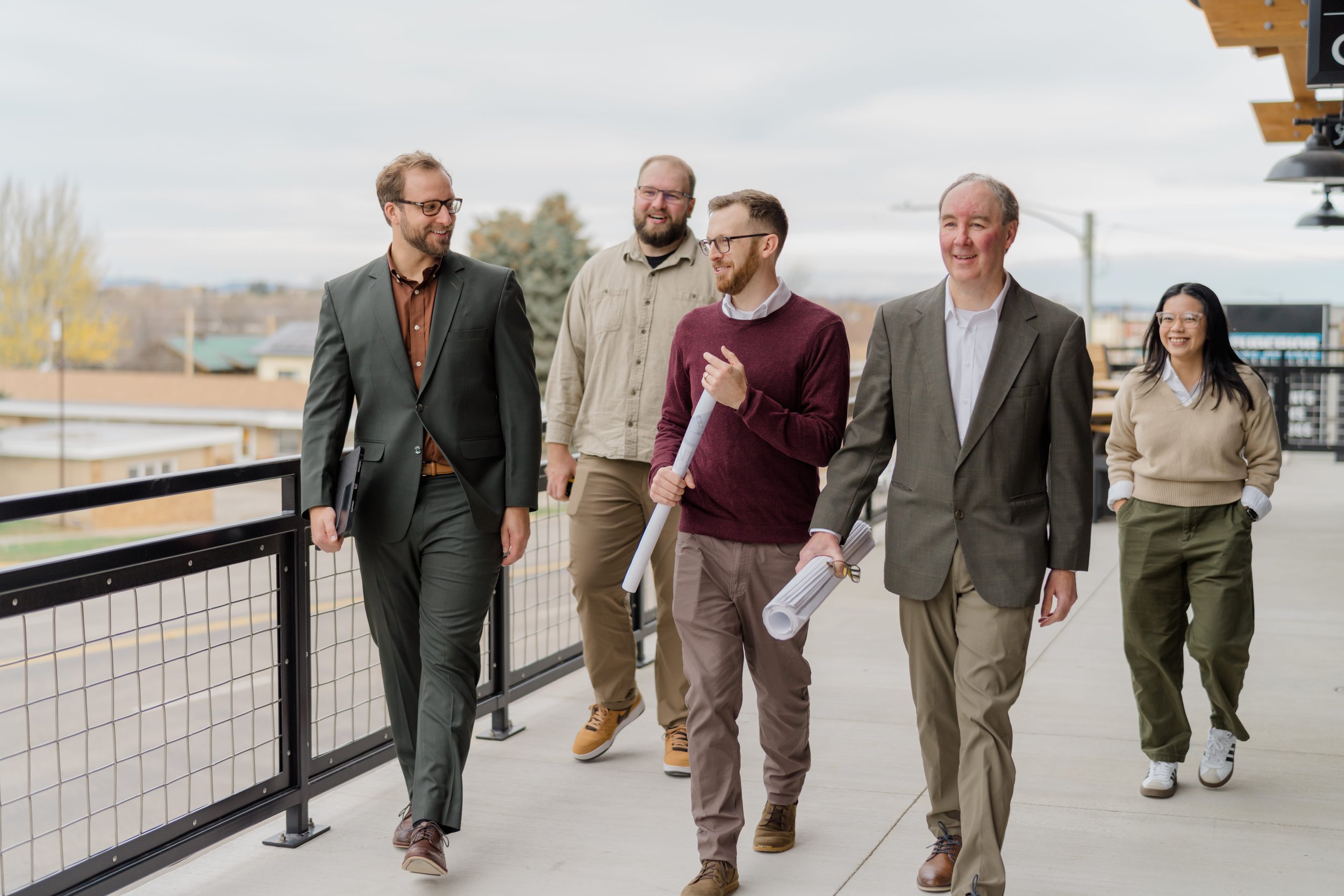 Group of six professionals walking and talking outside on a balcony with Maryland in background, some carrying blueprints.
