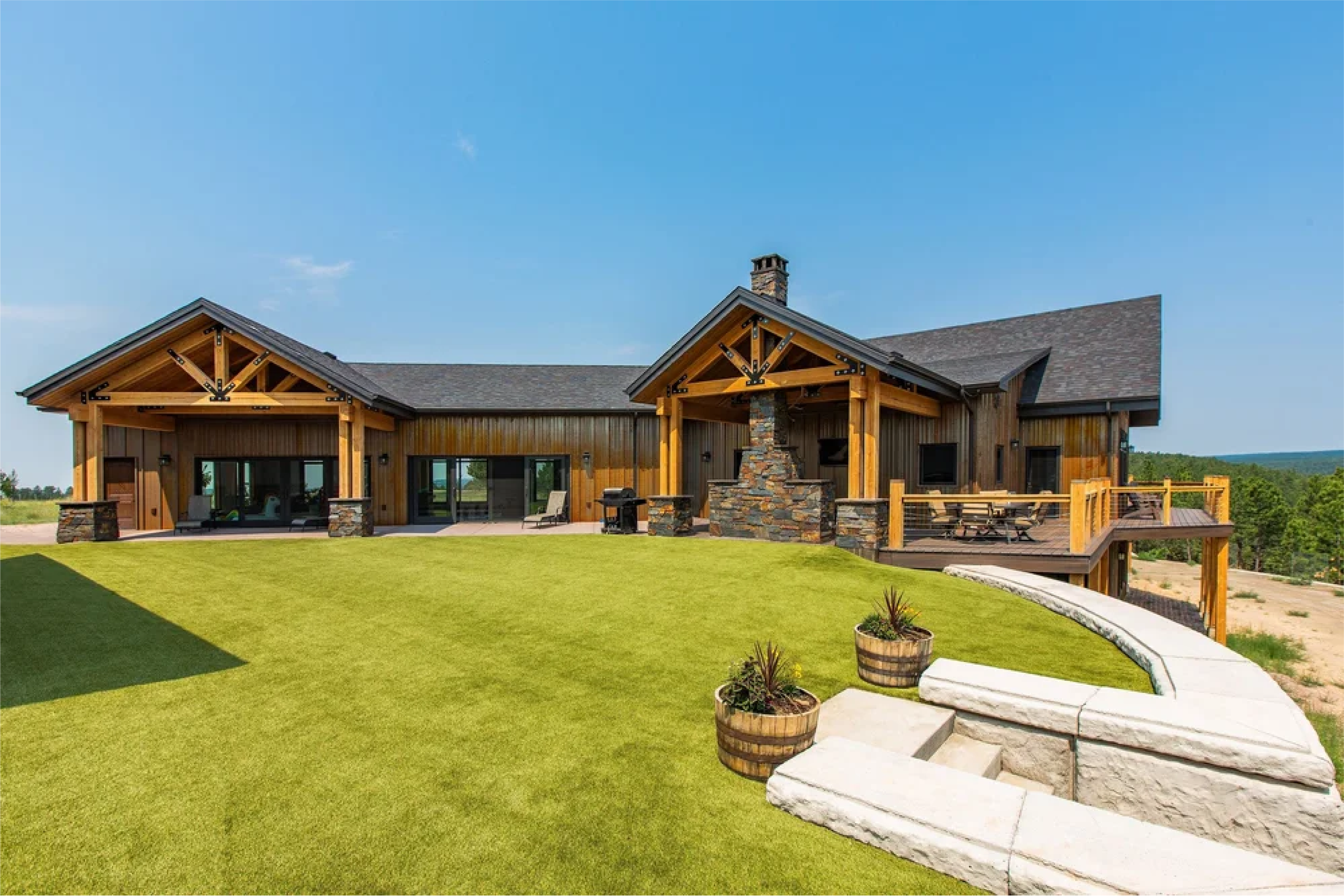 Large modern house with wooden exterior, stone accents, and a spacious deck overlooking a green lawn and distant trees.