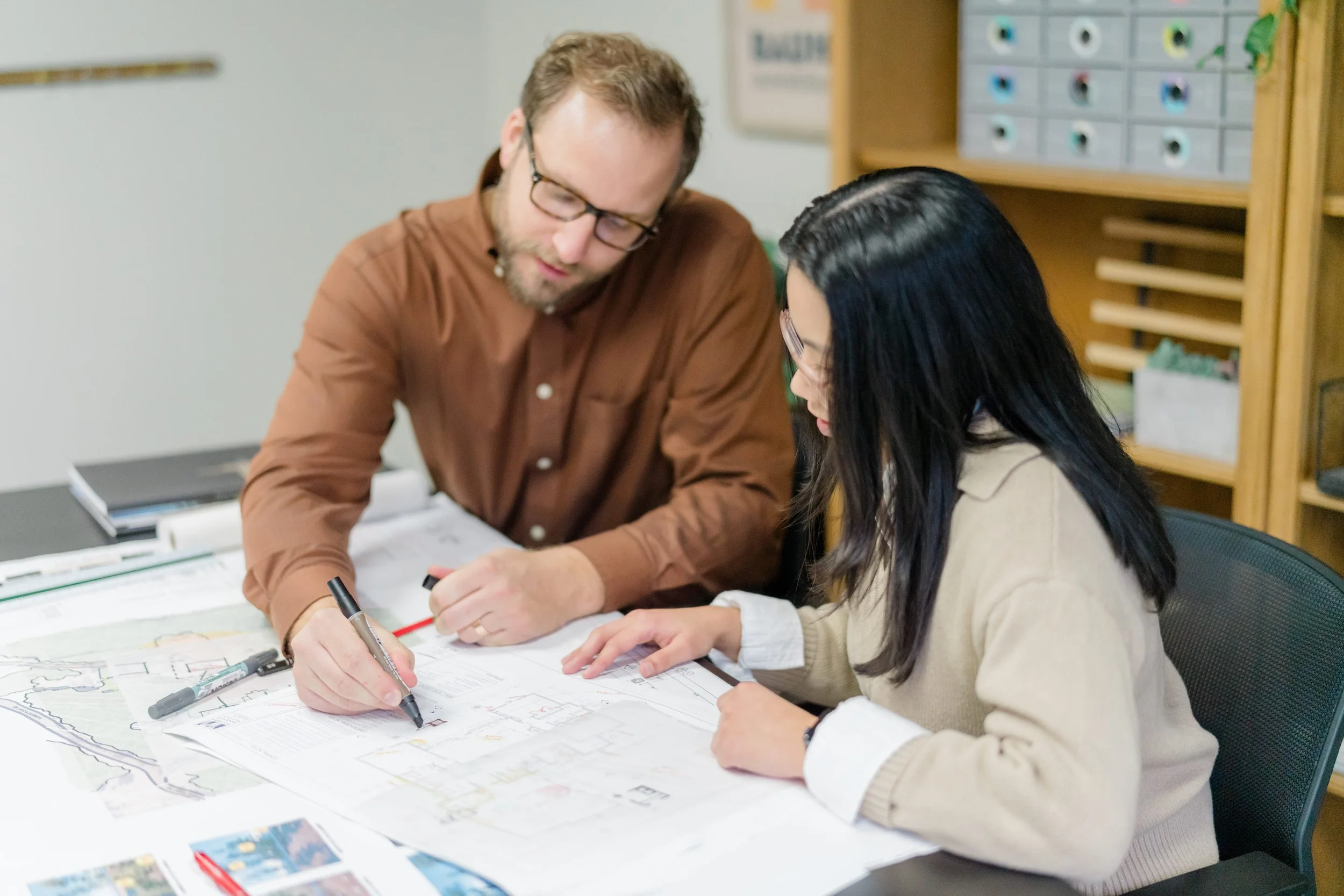 Two people, a man and a woman, working together at a table with architectural or engineering drawings, pens, and markers, in an office or classroom setting.
