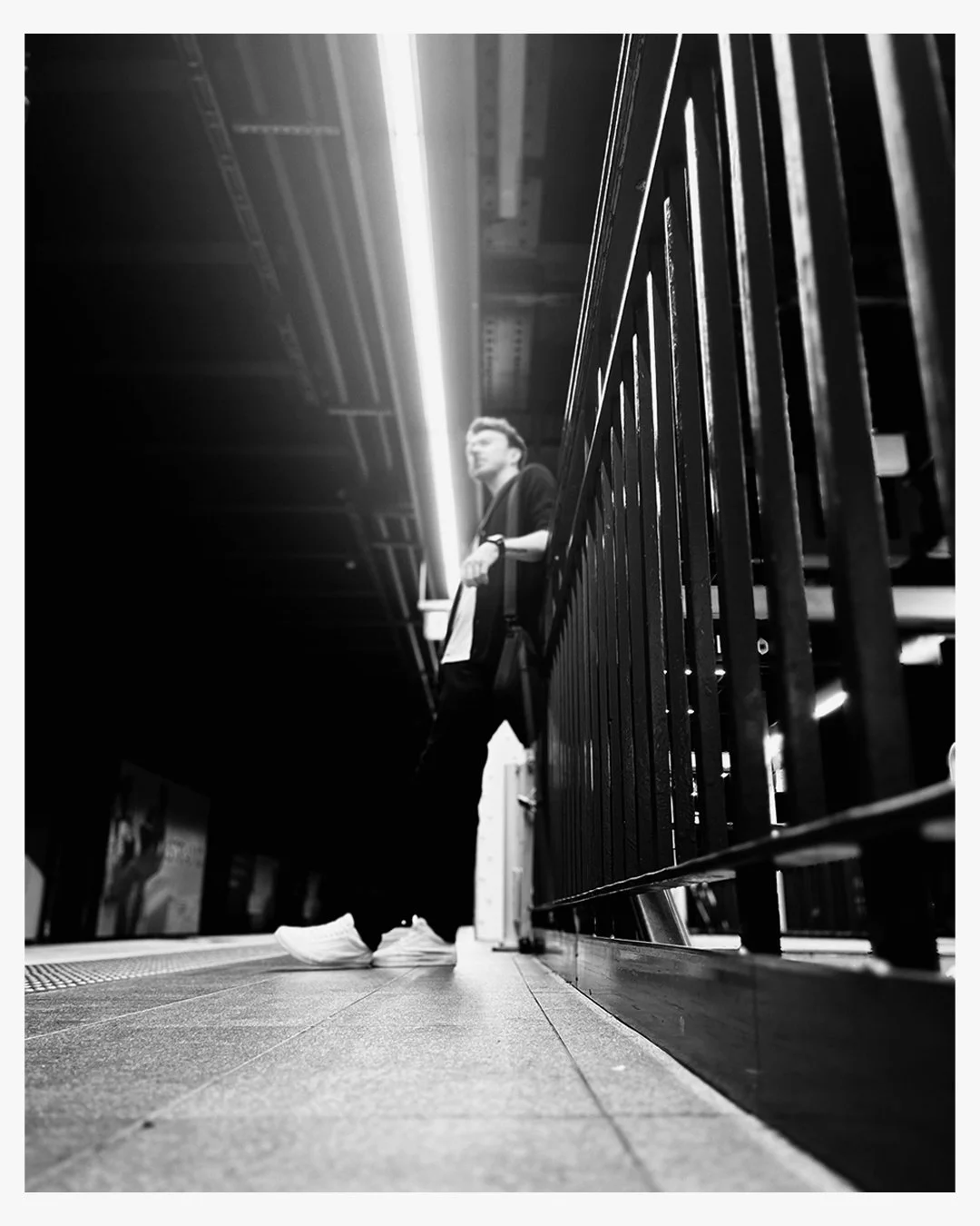 A black-and-white photo of a person standing on a tiled floor near a metal railing in an indoor space, possibly a subway or train station, illuminated by a long bright light fixture on the ceiling.