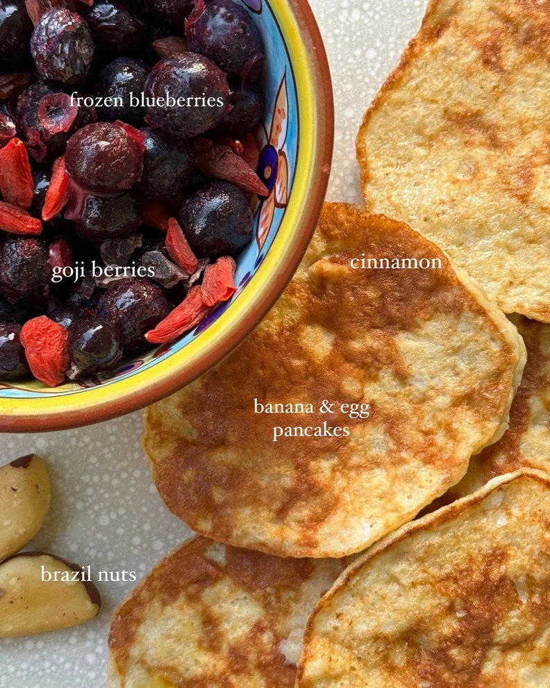 Bowl of frozen blueberries and goji berries next to banana and egg pancakes topped with cinnamon. Small portion of Brazil nuts are also visible.