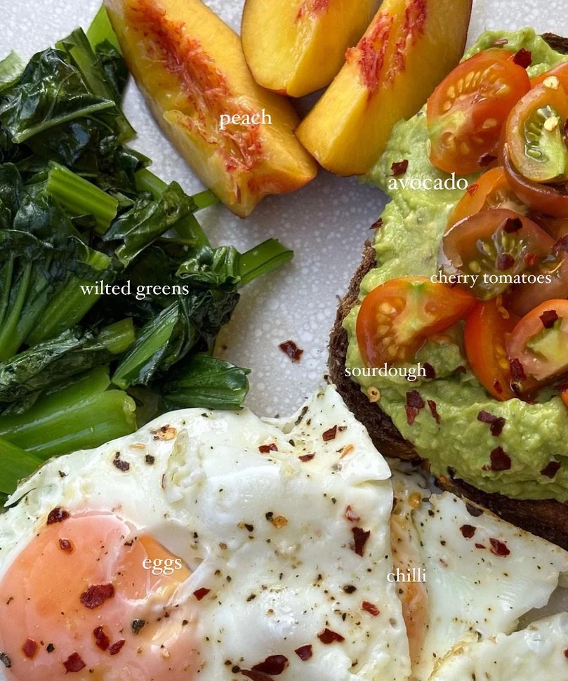 A breakfast plate with peach slices, wilted greens, avocado toast topped with cherry tomatoes, sourdough bread, eggs, and a sprinkle of chili flakes.