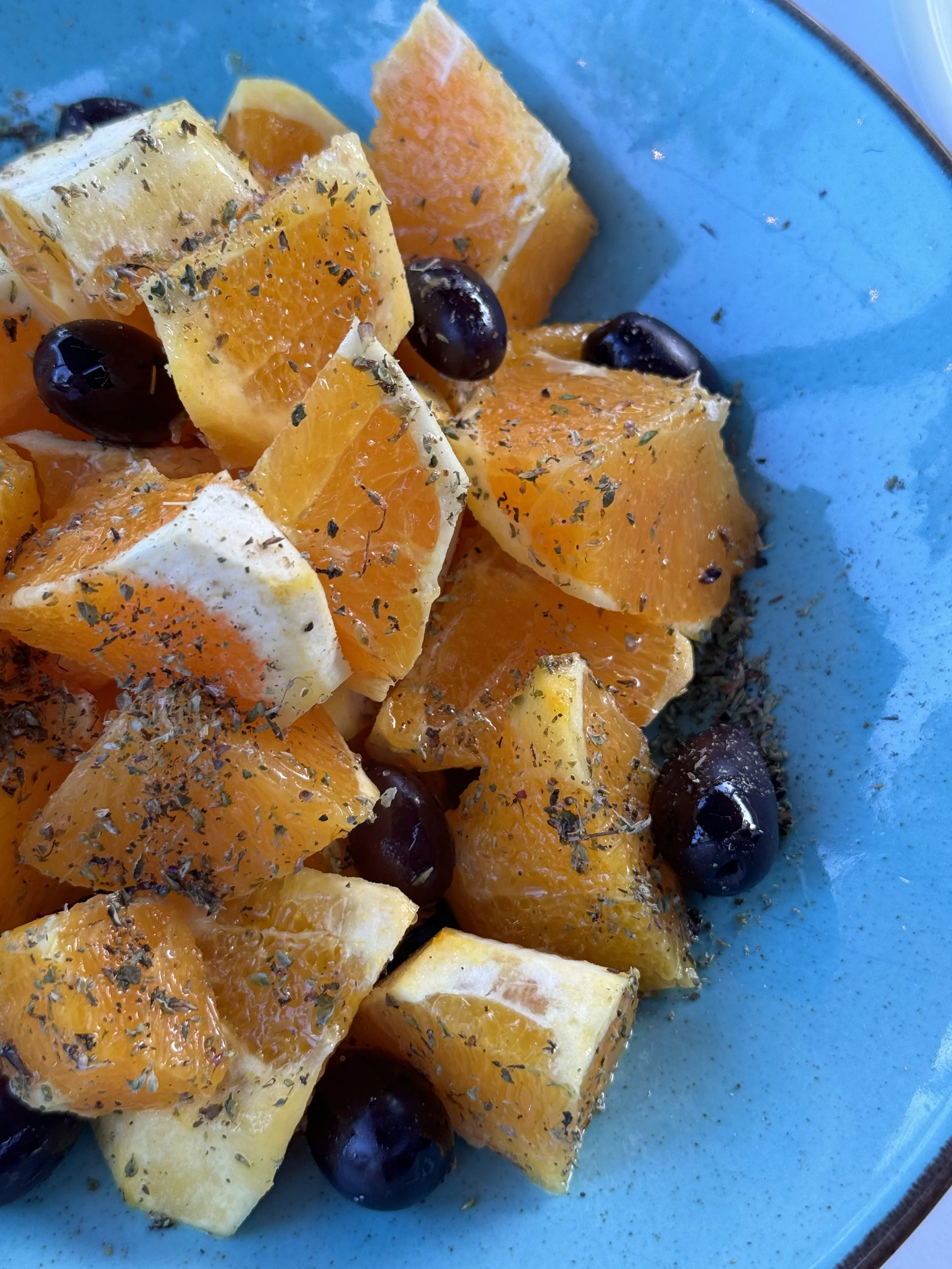 A salad with orange segments, pineapple chunks, black olives, and herbs on a blue plate.
