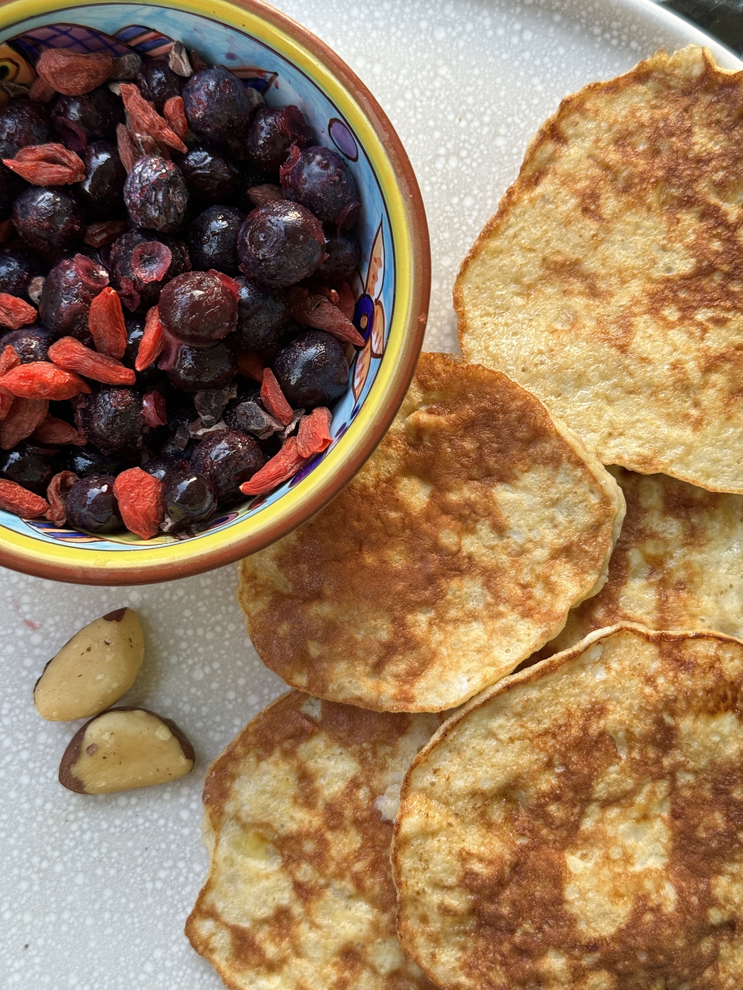 A plate with four fried potato pancakes, a bowl of blueberries topped with goji berries, and two almonds.