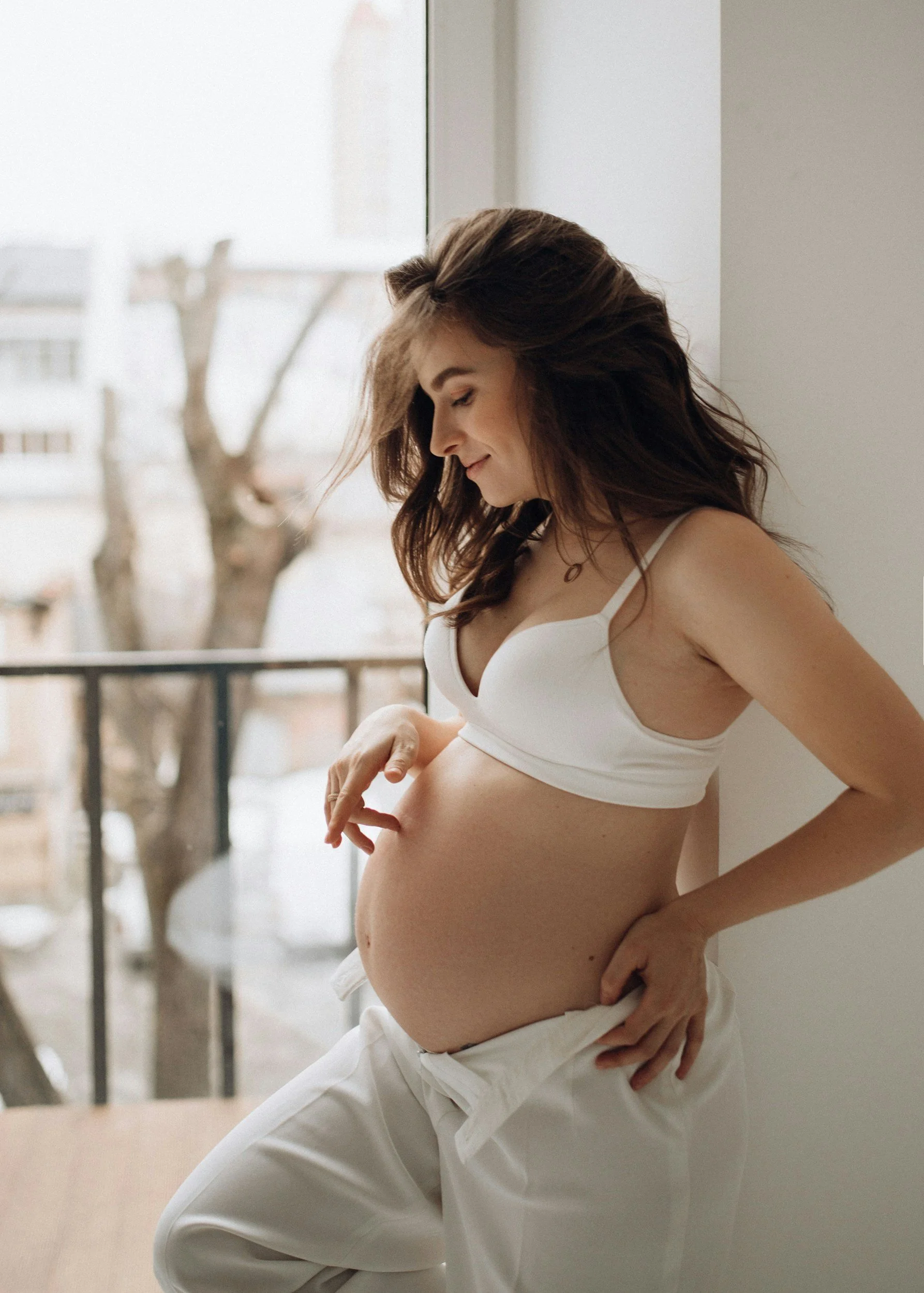 A pregnant woman with brown hair in white clothing looking and touching her baby bump indoors near a window.